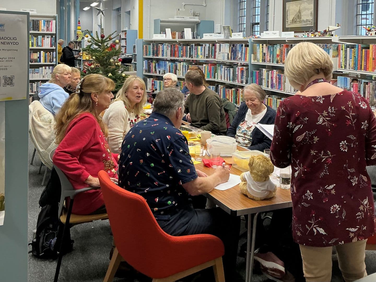 A group of people sit around a table in a library, chatting and sharing food near a decorated Christmas tree. - Home Instead