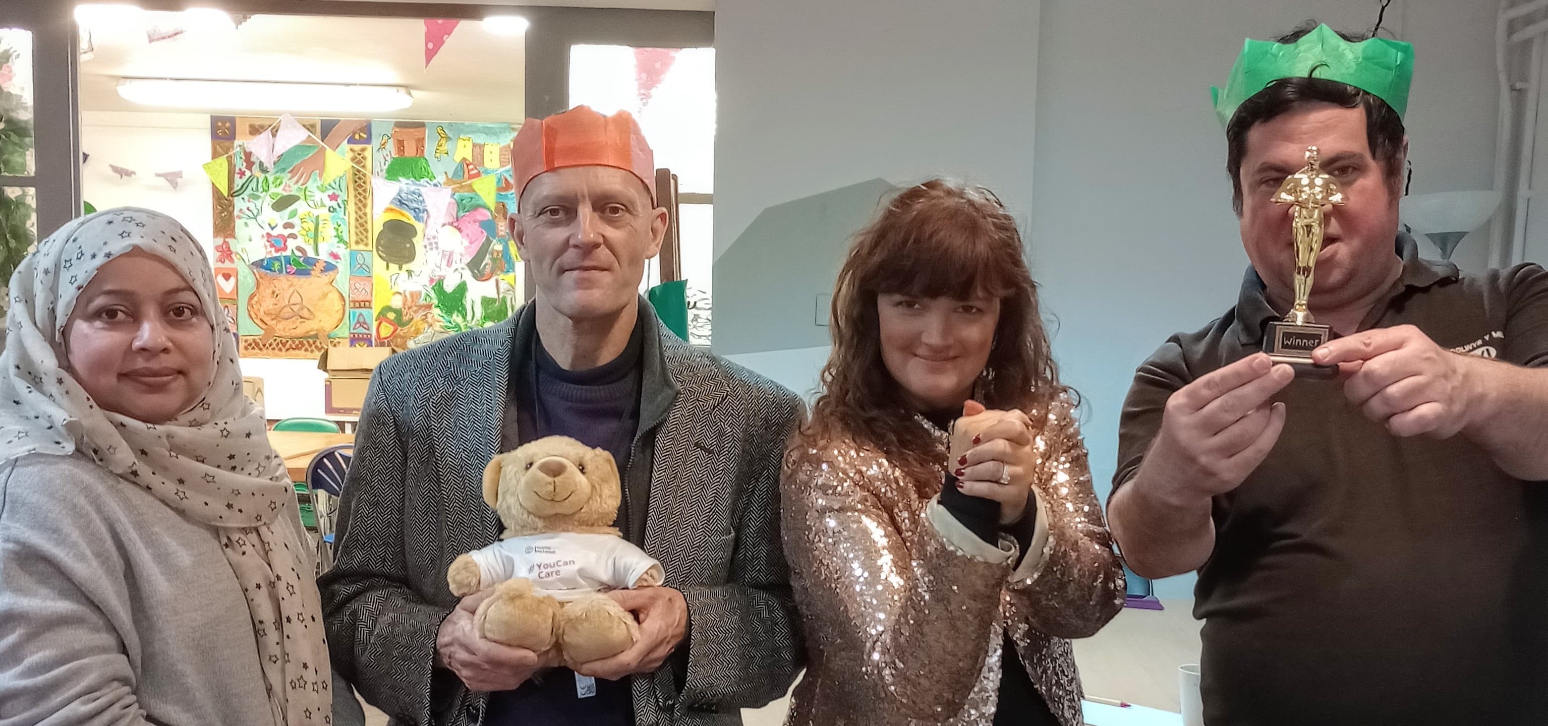 Four people in festive hats, one holding a teddy bear, another holding a small trophy, posing and smiling. - Home Instead