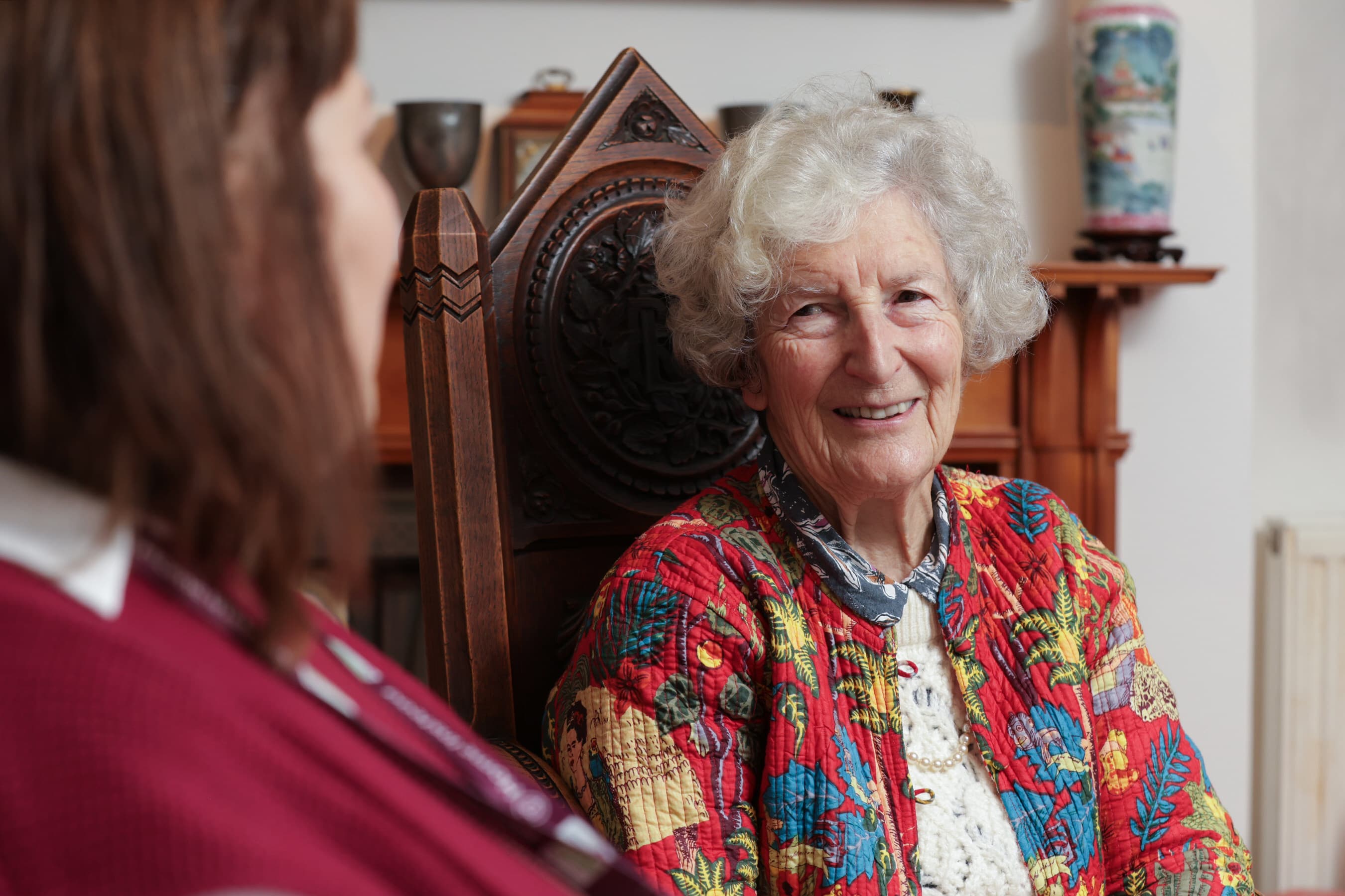 Elderly woman in a colorful jacket smiles while talking to another person indoors. - Home Instead