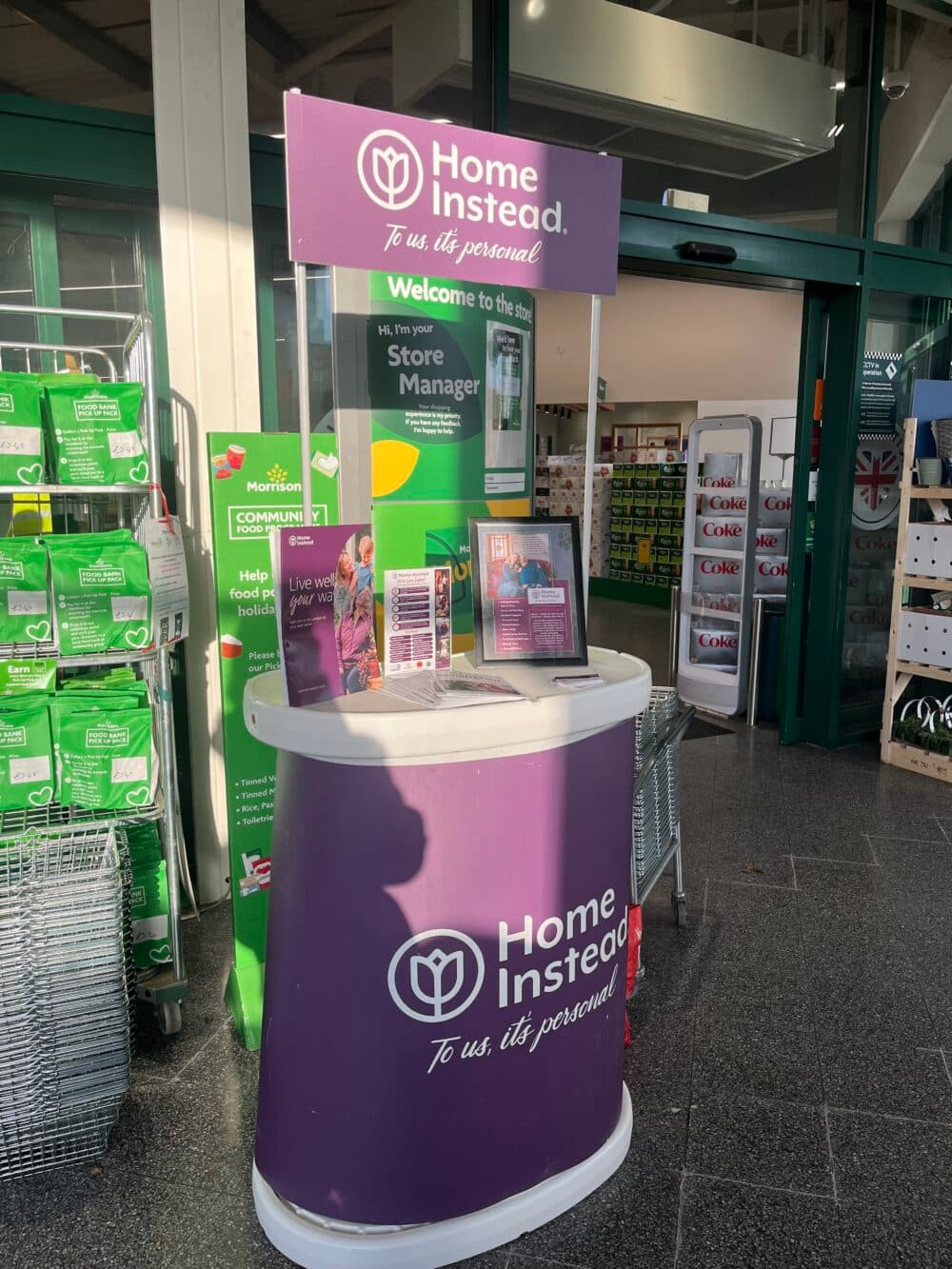 A "Home Instead" information booth stands inside a store entrance, with brochures and a framed sign displayed. - Home Instead
