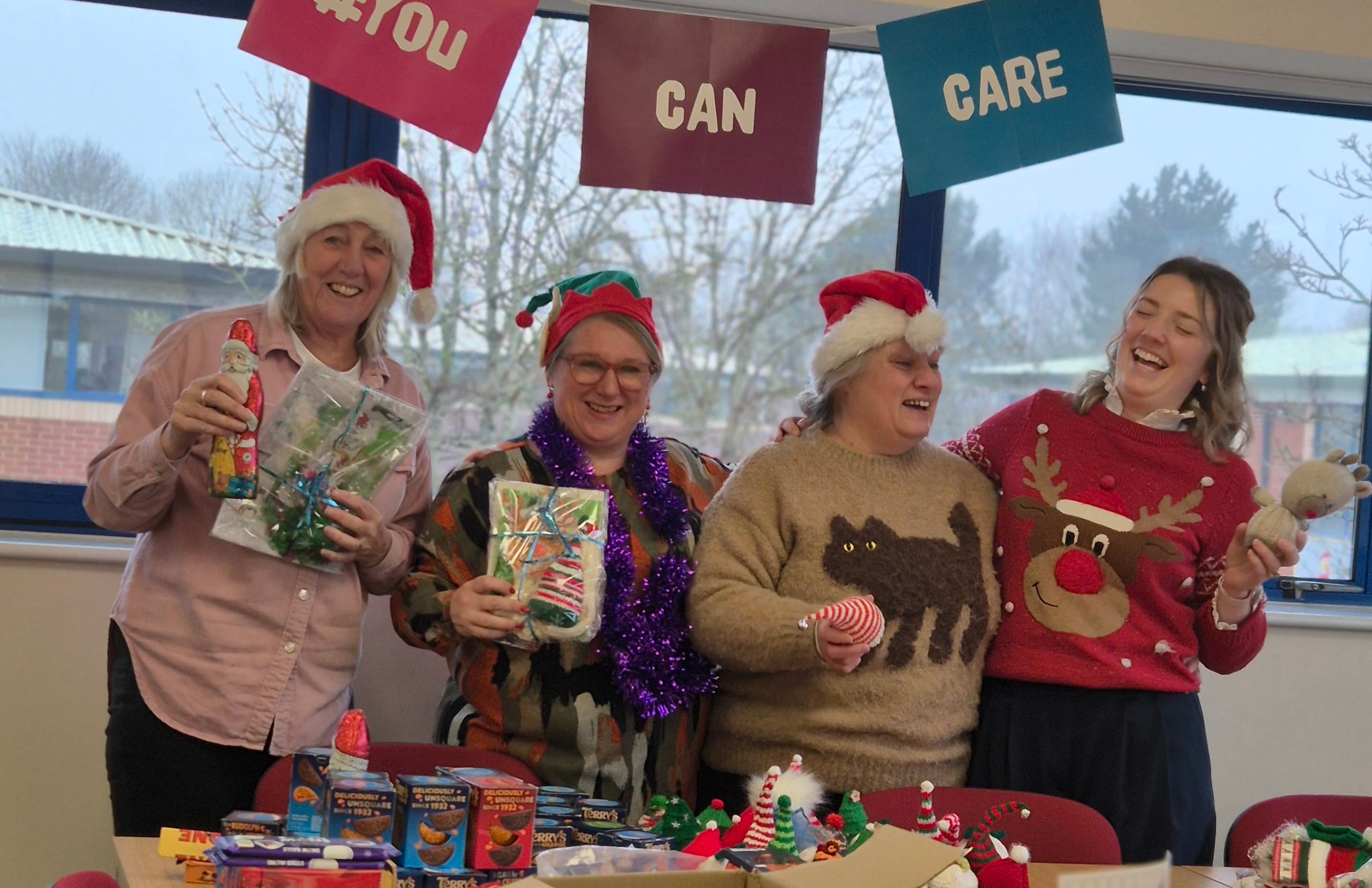 Four people in festive sweaters and hats smile holding gifts, standing by a table of donated food and supplies. - Home Instead