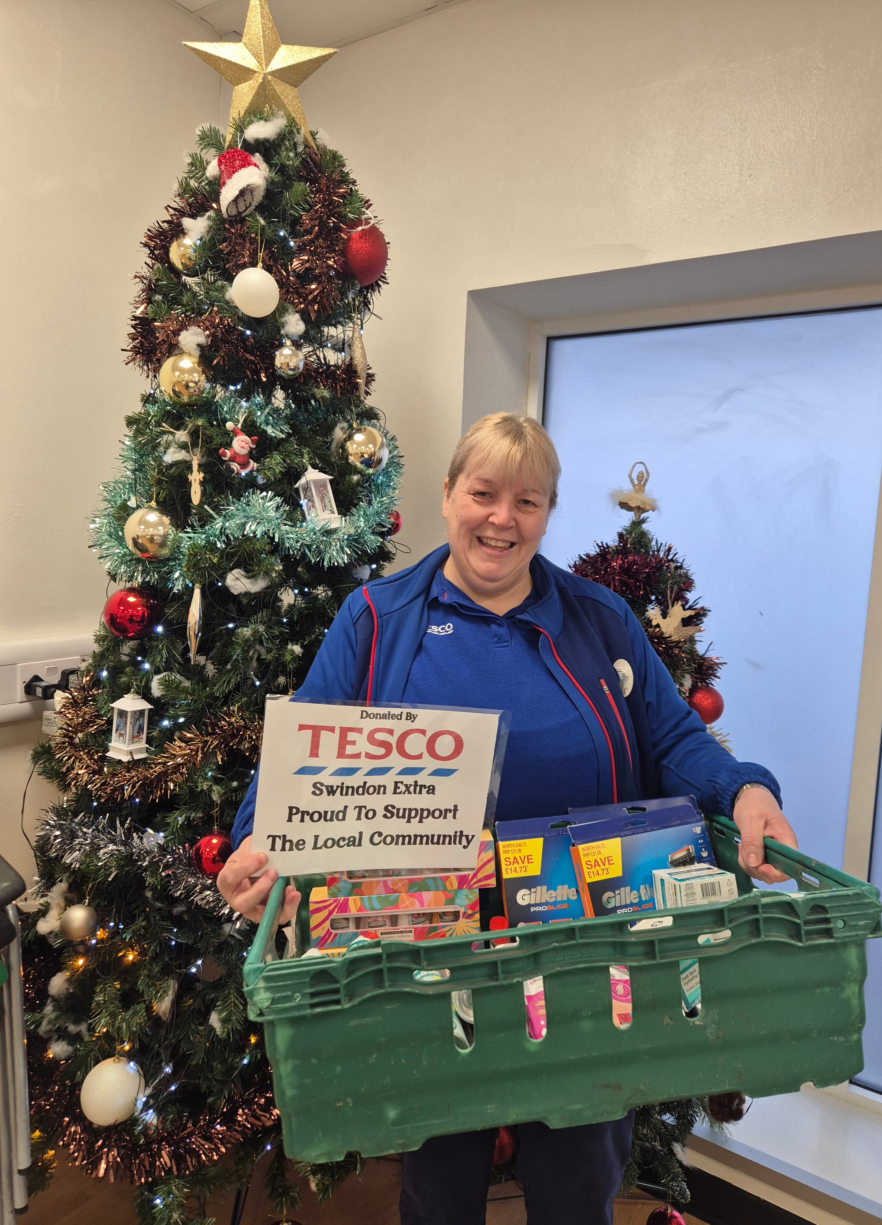 Smiling woman by a Christmas tree holds a basket of donations and a Tesco sign supporting the local community. - Home Instead
