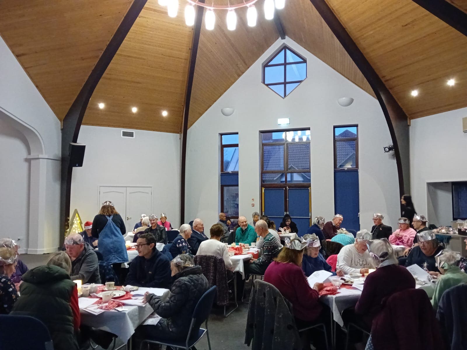People wearing party hats sit at decorated tables in a hall with a wooden ceiling, enjoying a festive meal. - Home Instead
