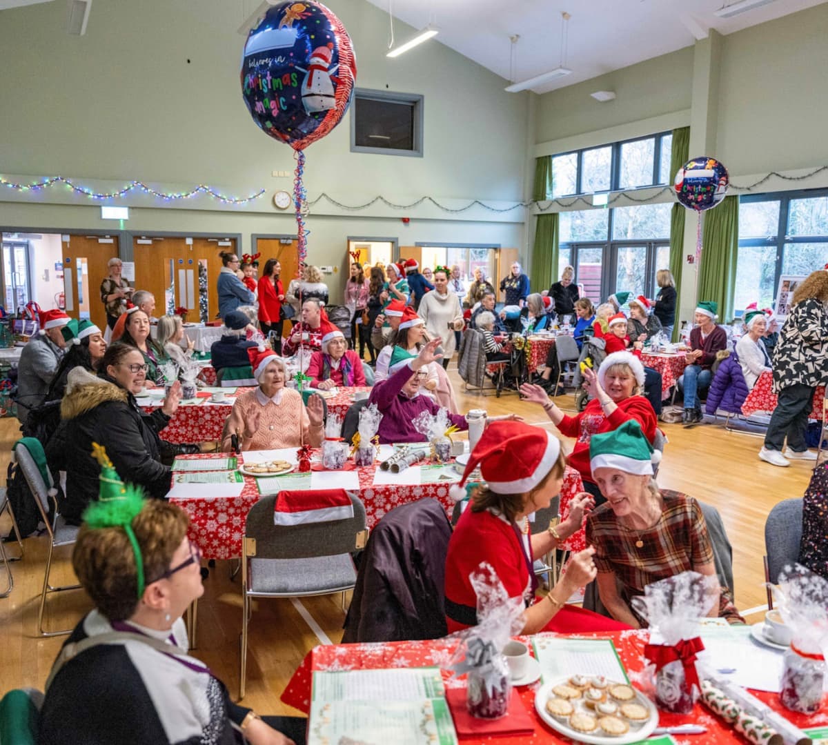 People having a prty with balloons and hats and long tables and some are wearing red and green clothes and all are happy and smiling inside the hall