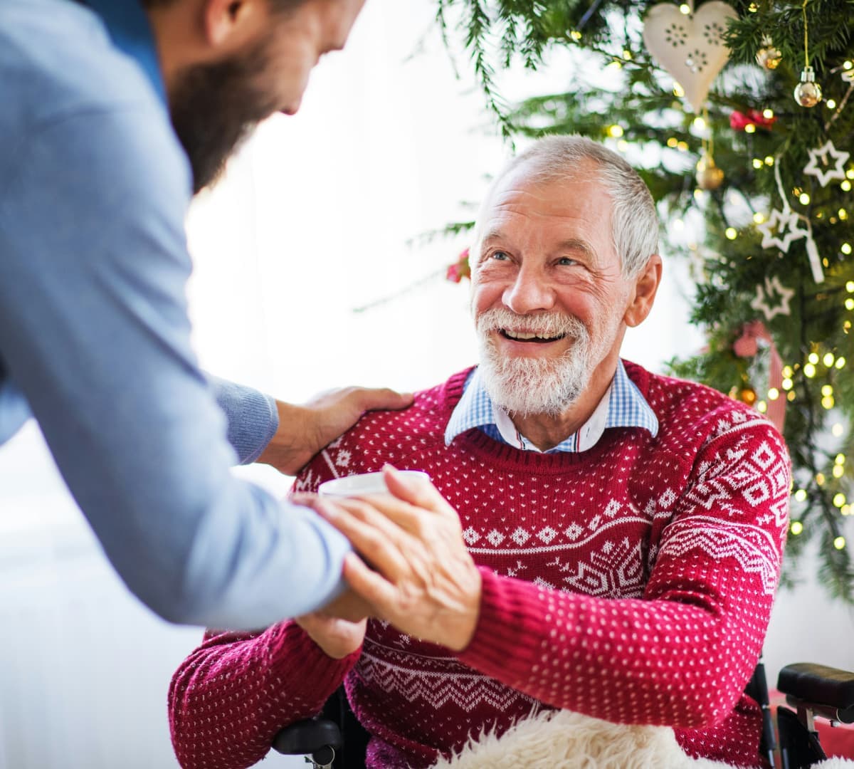 Senior man with grey hair and moustache and smiling and wearing red sweatshirt while sitting on a wheelchair and shaking the hand of his son and with Christmas tree at the background