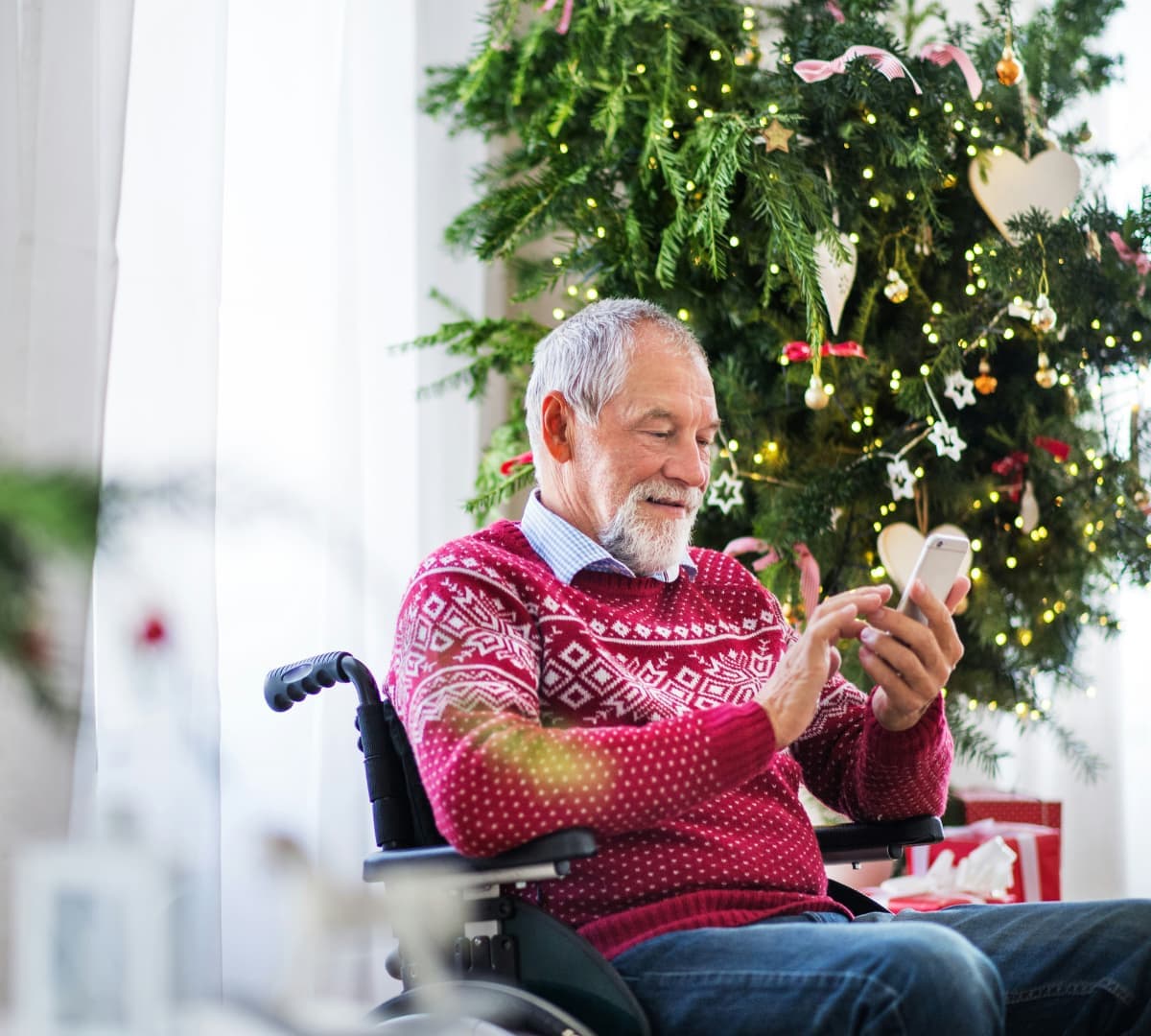Senior man sitting on a wheelchair wearing a red sweater and using his phone while smiling with Christmas tree on the background