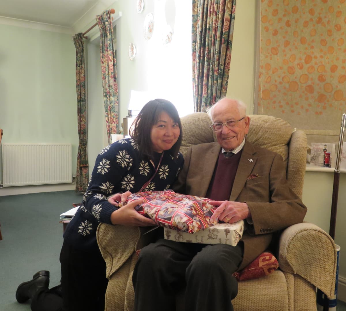 Senior man sitting on a brown couch while holding a gift with his carer kneeling at the floor inside the home