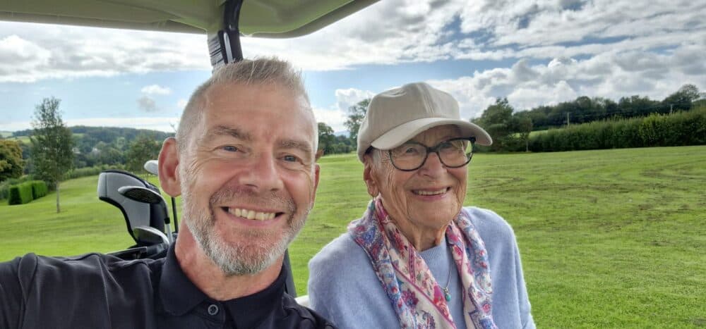 Smiling man and elderly woman wearing a hat sit together in a golf cart on a grassy field. - Home Instead