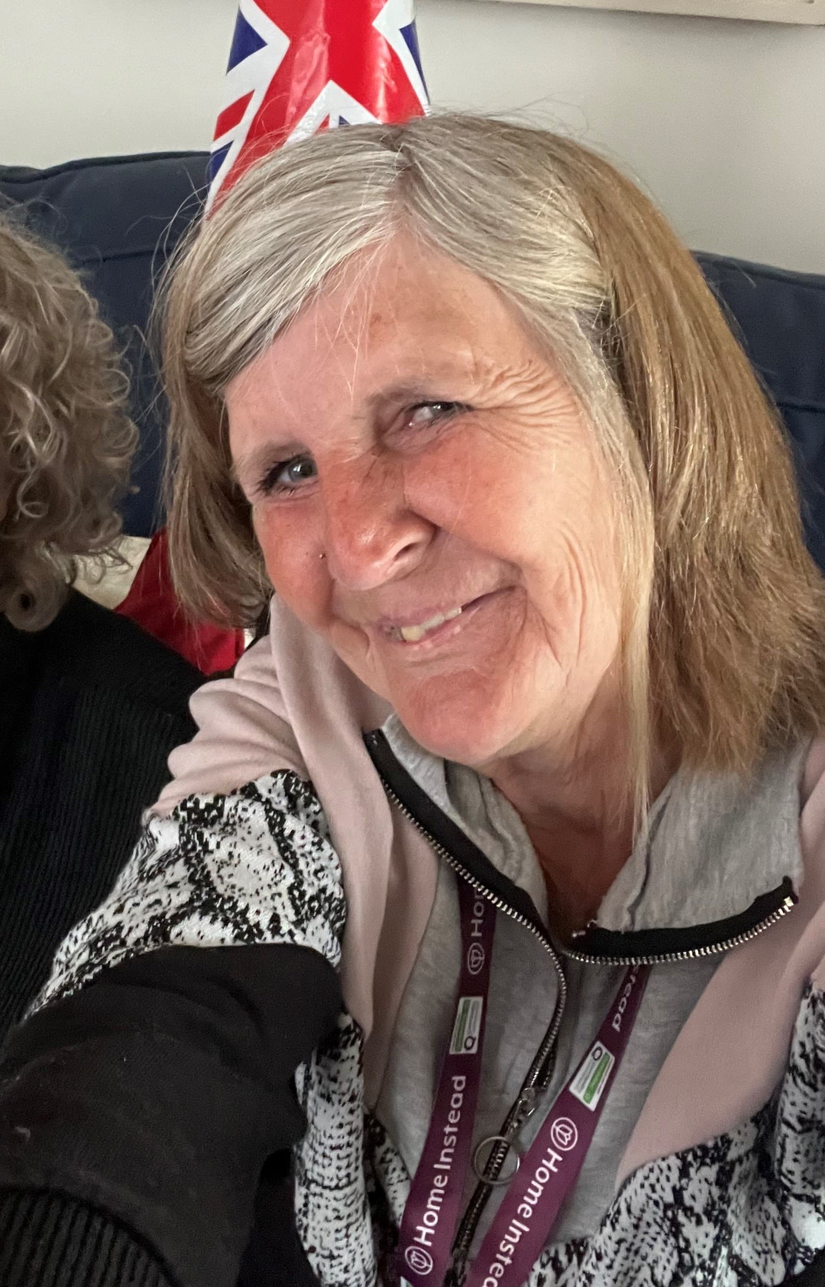 Smiling older woman with gray hair and lanyard, sitting indoors with a Union Jack flag behind her. - Home Instead