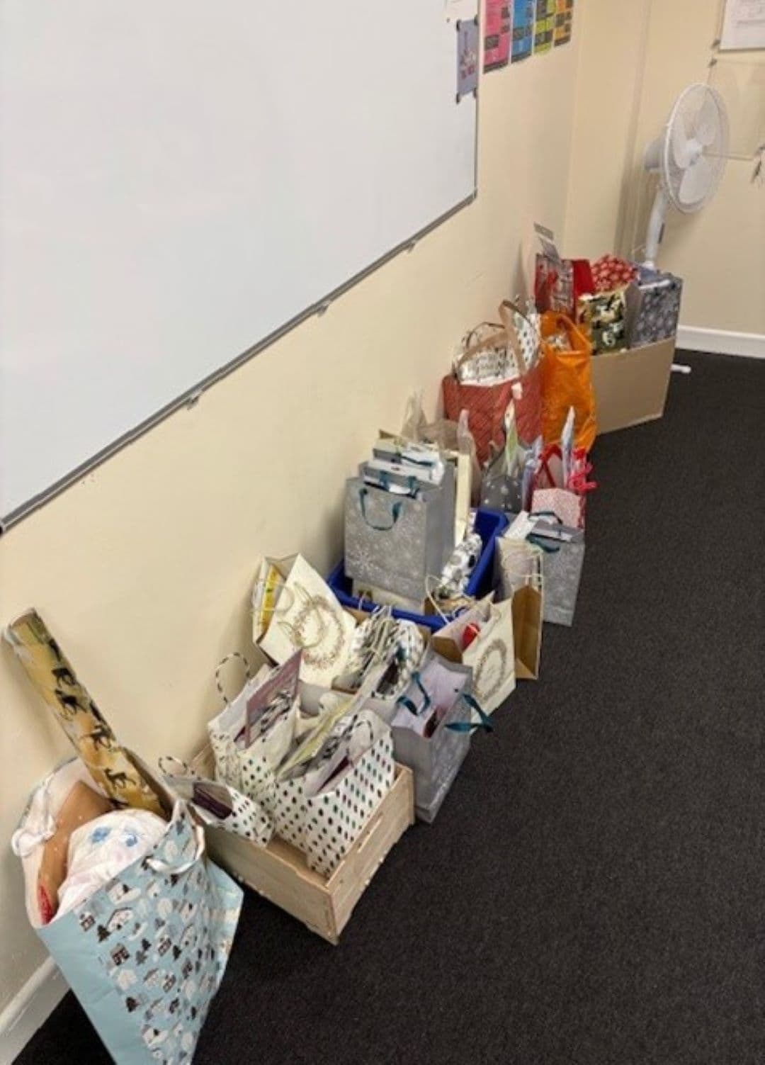 A row of gift bags and presents lined up on the floor against a classroom wall. - Home Instead
