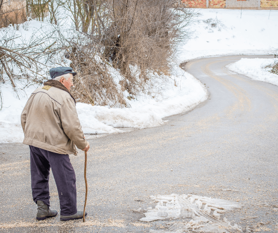 Elderly man walking down a snowy road with a cane. He is wearing a brown coat and a blue hat walking cautiously.
