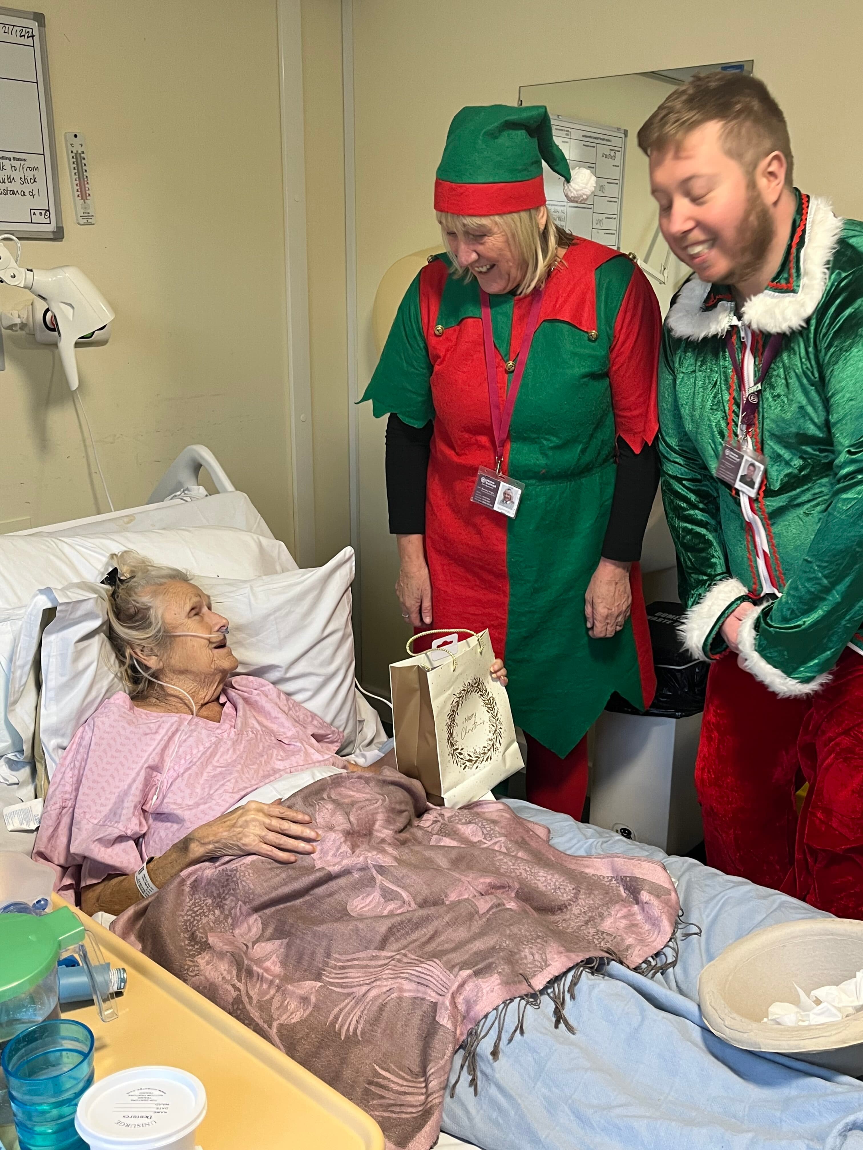 An elderly woman in a hospital bed smiles at two people dressed as Christmas elves giving her a gift bag. - Home Instead