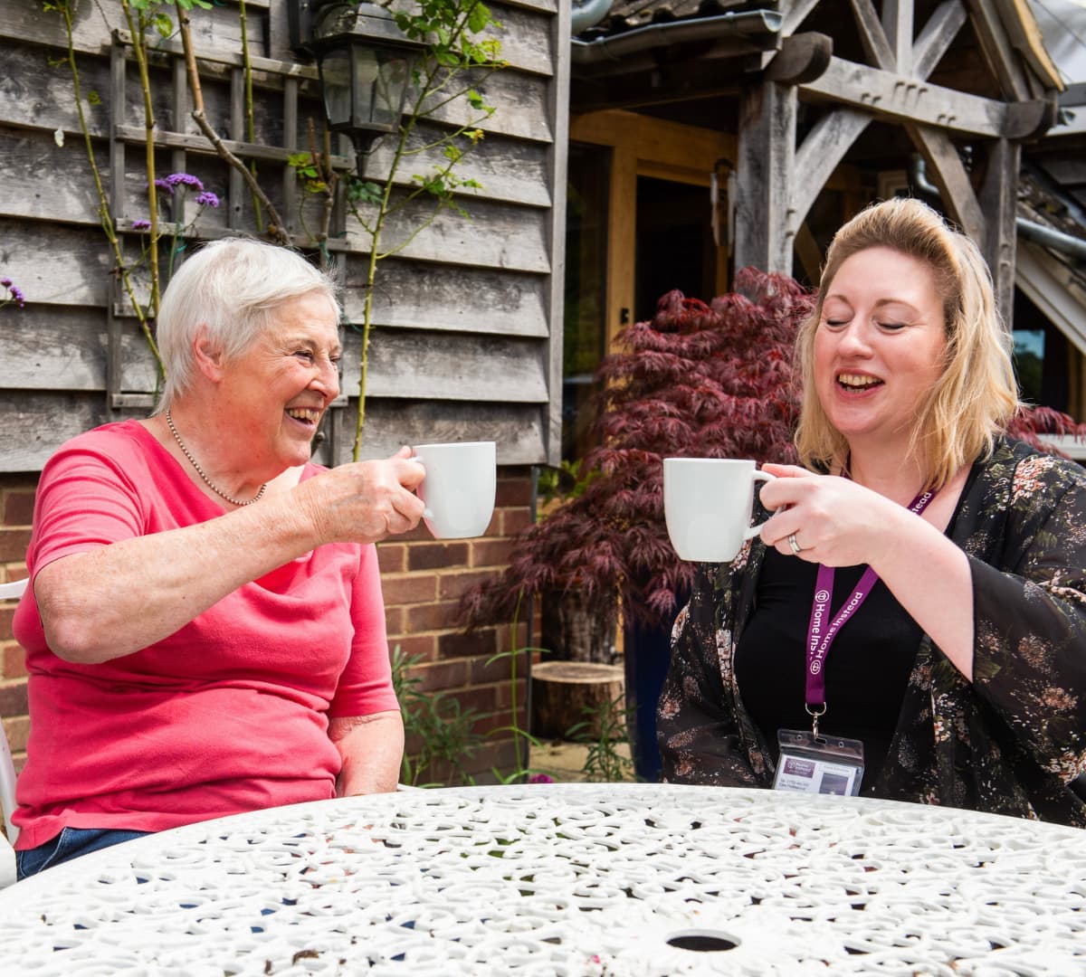 An older adult with short grey hair drinking coffee using white mugs outside of her house with her carer both happy and smiling