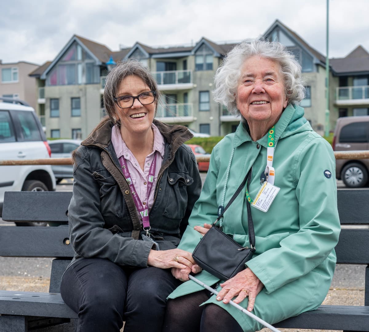 Older woman with grey short hair and wearing green while sitting on a bench and enjoying the outdoor with her carer