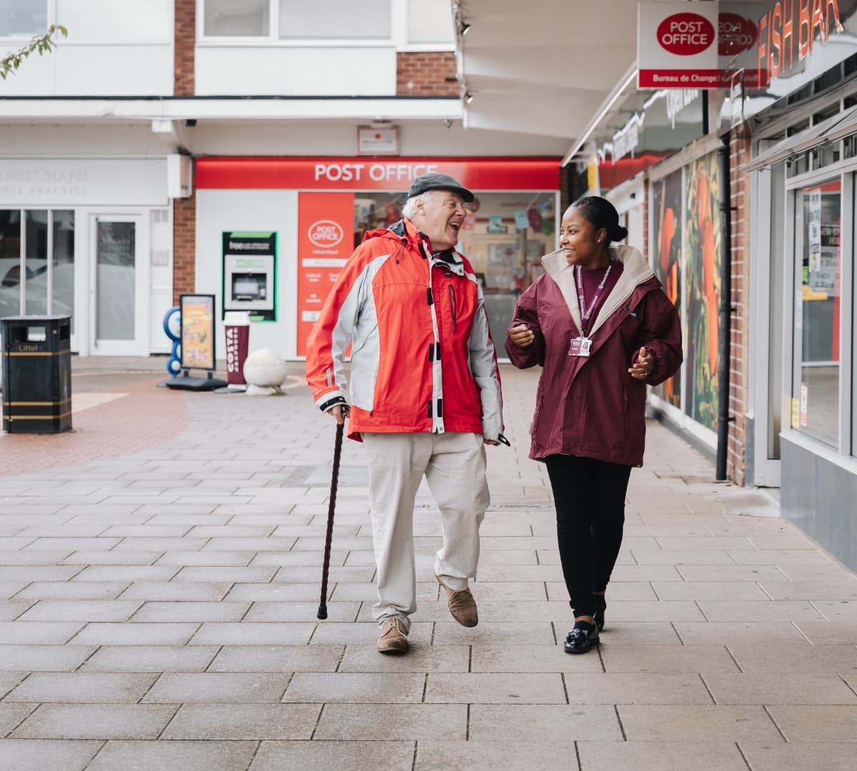 A senior man with a crutch walking with his carer in a cold afternoon with different establishments around