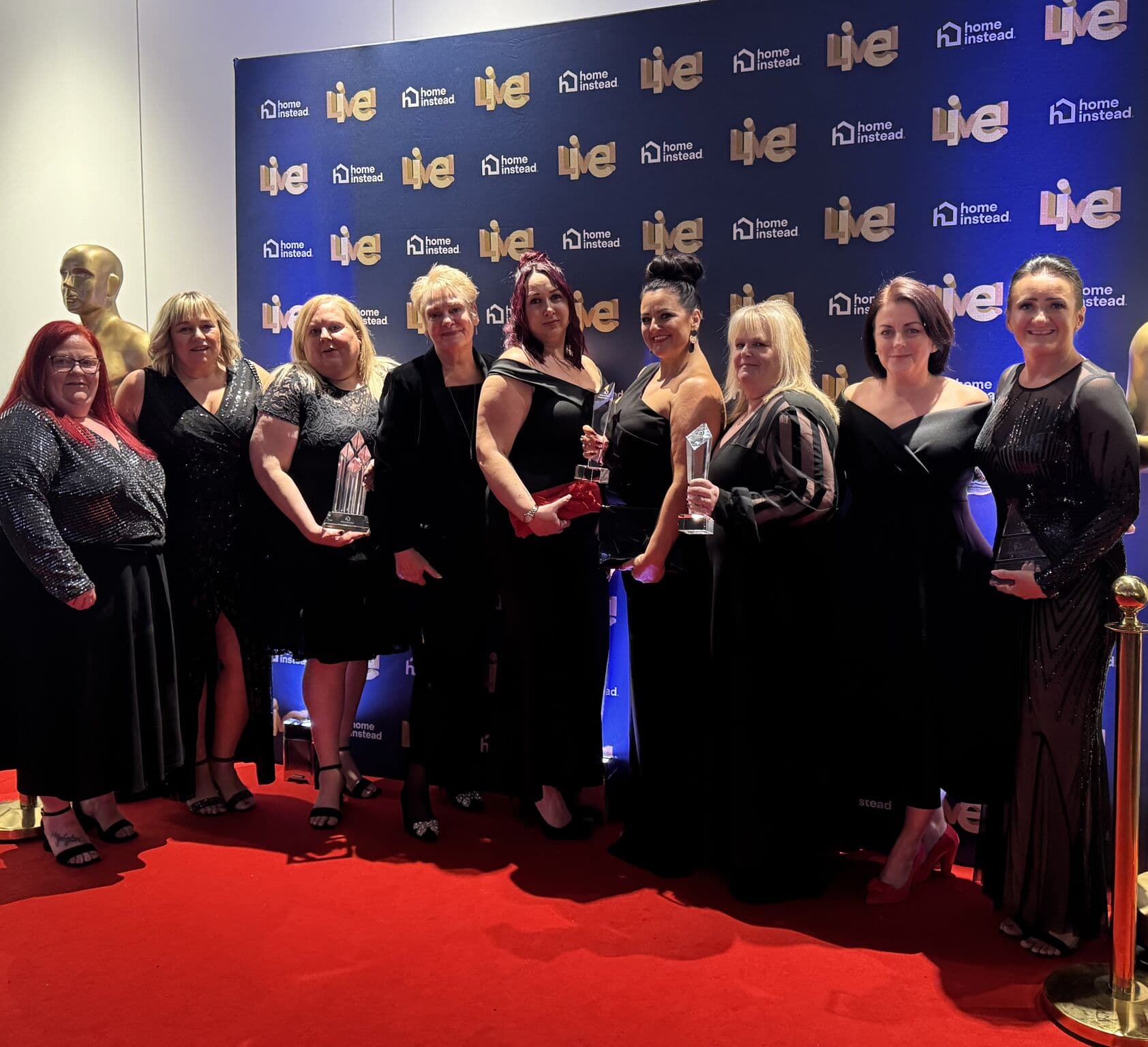 Ten women in formal dresses pose with trophies on a red carpet in front of a branded event backdrop. - Home Instead