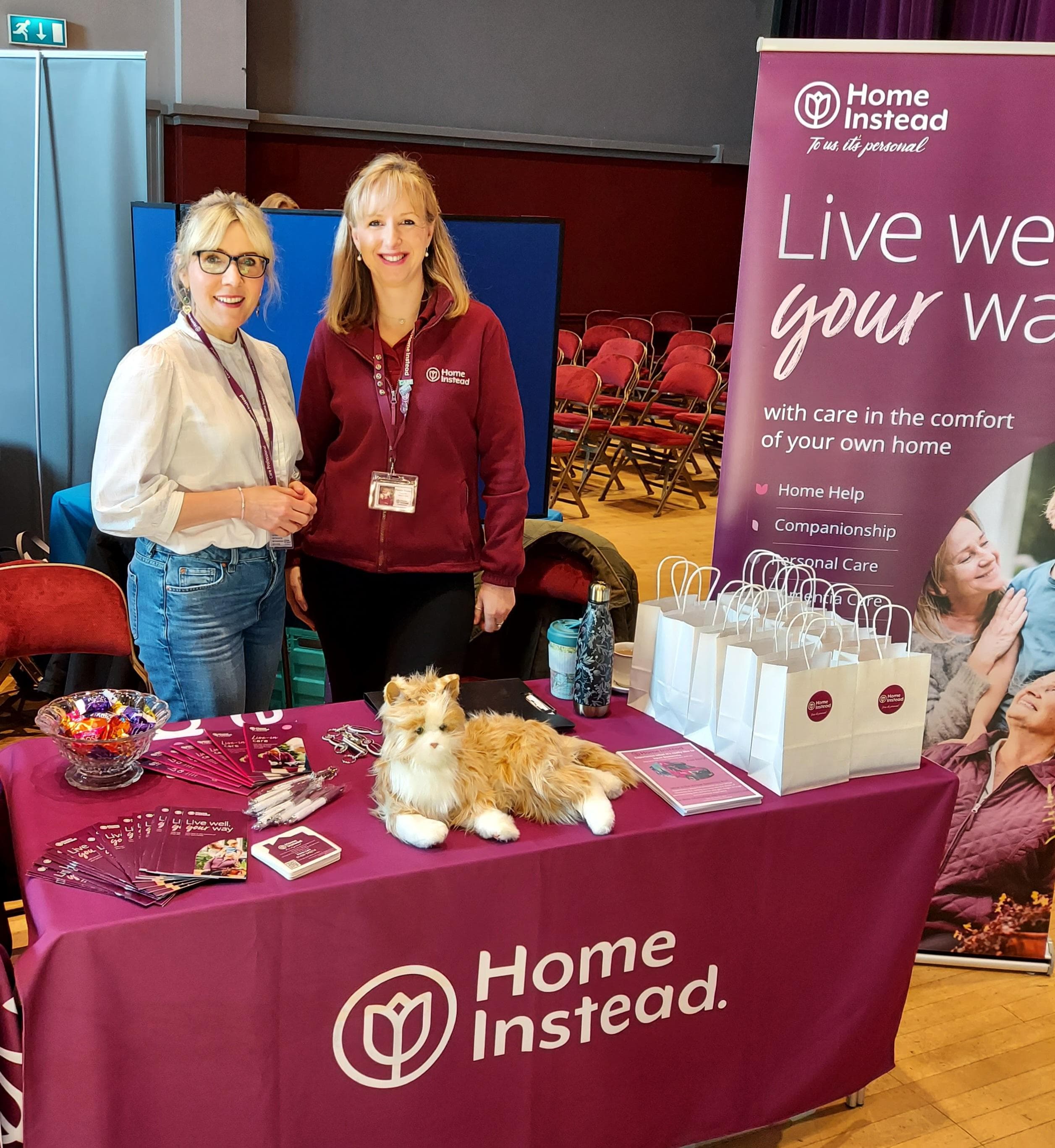 Two women stand at a Home Instead booth with brochures, gift bags, and a robotic cat on the table. - Home Instead