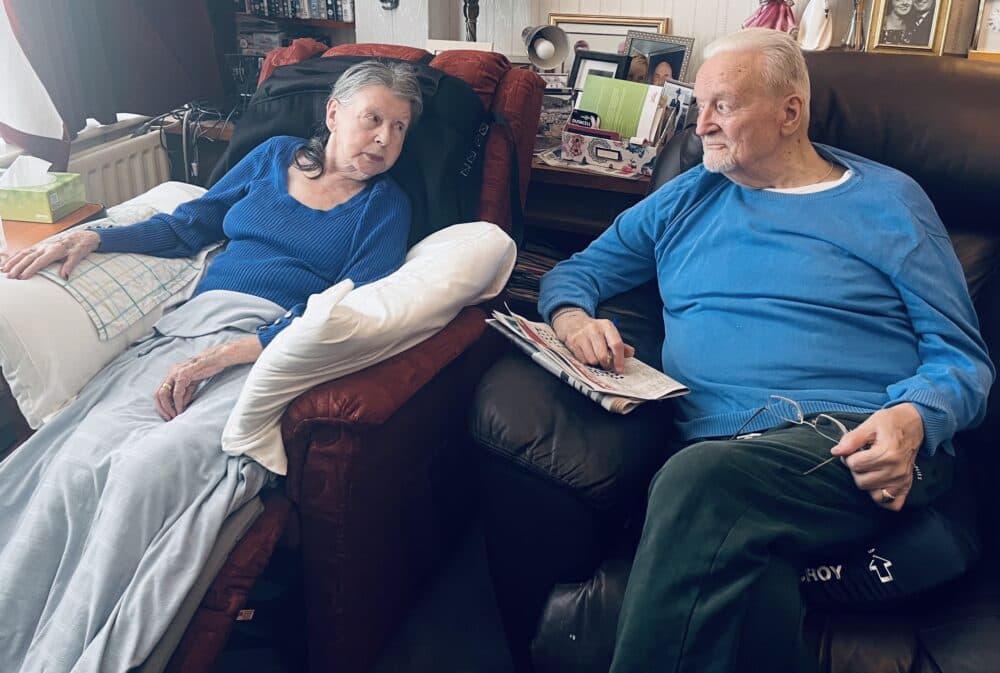 Elderly woman reclines on a chair while an elderly man sits nearby holding a newspaper; both look at each other. - Home Instead