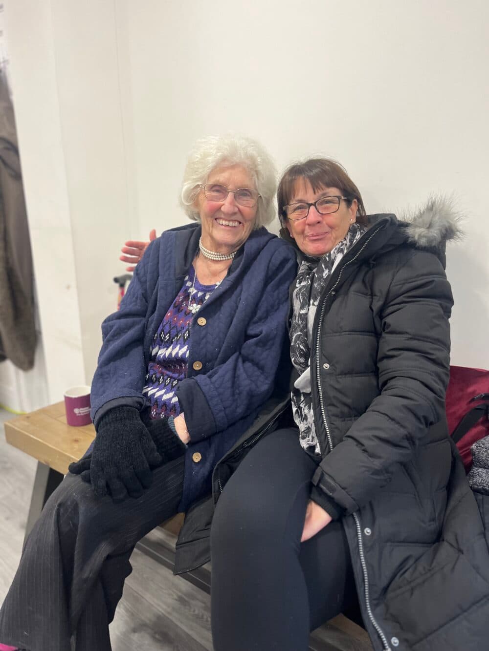 Two women sit close together on a bench, smiling warmly at the camera in a cozy indoor setting. - Home Instead