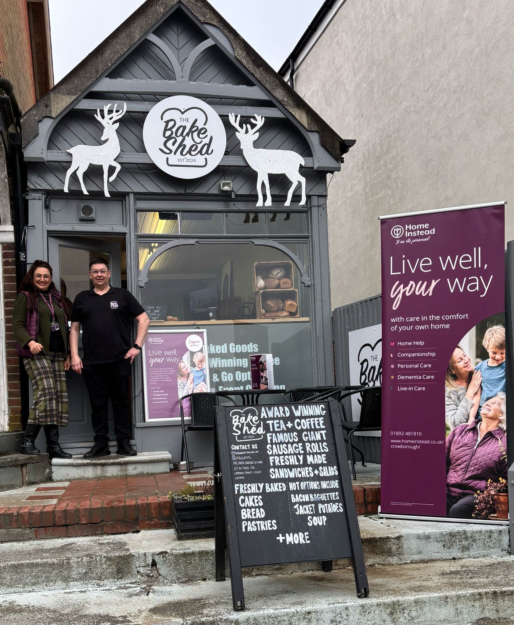 Two people stand outside The Bake Shed café, next to a signboard and a "Live well, your way" banner. - Home Instead