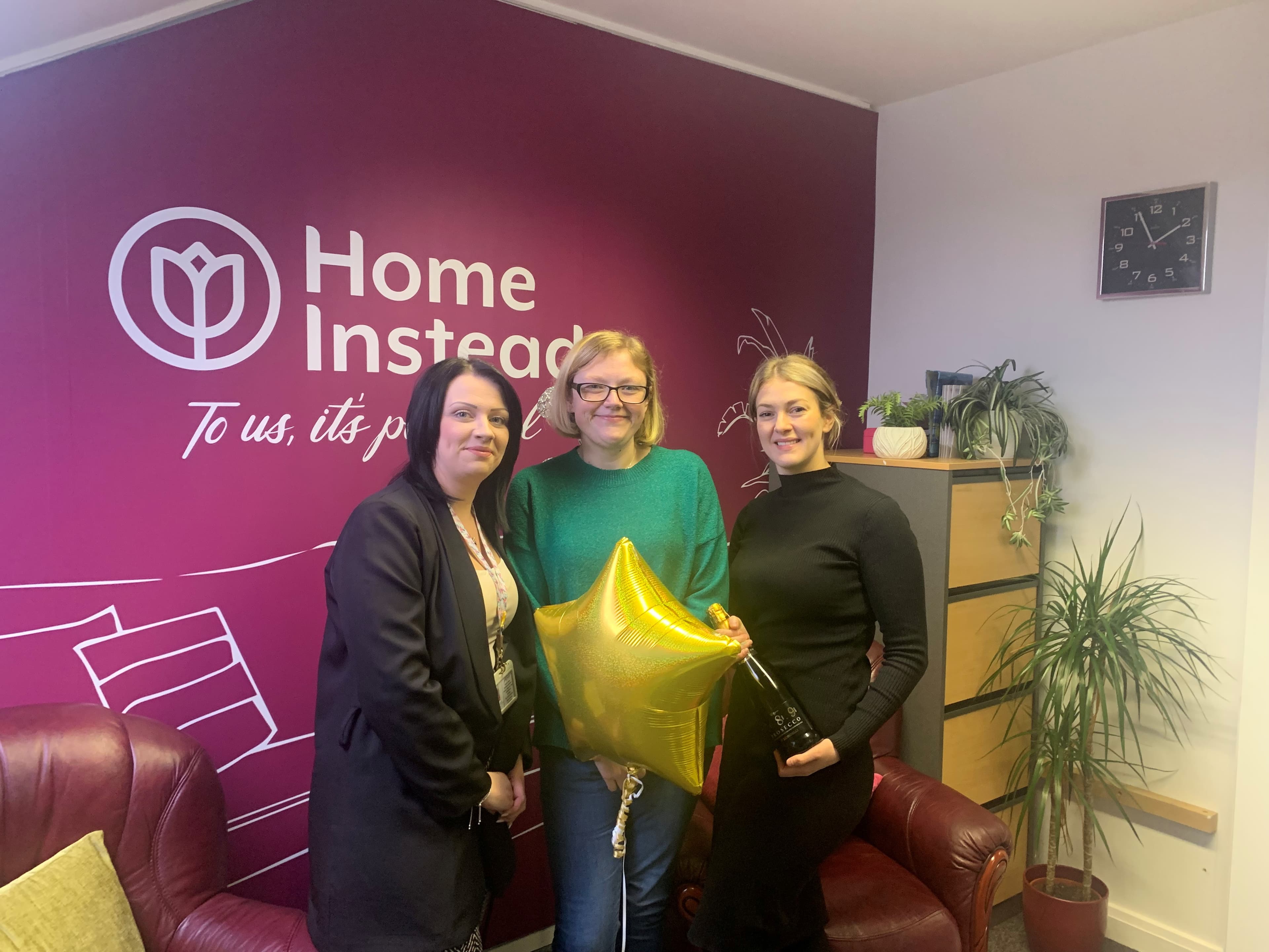 Three women smiling indoors by a burgundy Home Instead wall, holding a gold star balloon and a bottle. - Home Instead