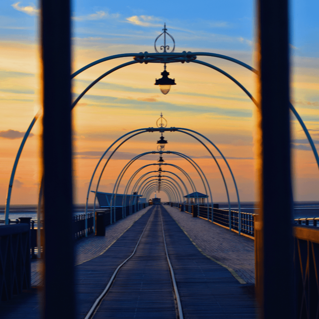 Sunset view of a pier with curved lamp posts, seen through railings, leading out over the water. - Home Instead