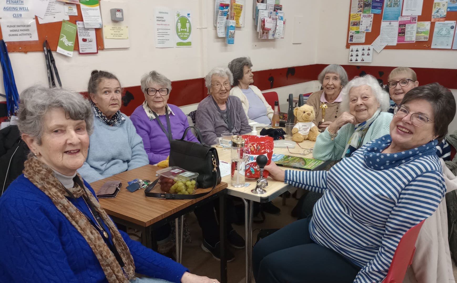 Eight older women sit smiling around a table at a social gathering in a community room. - Home Instead