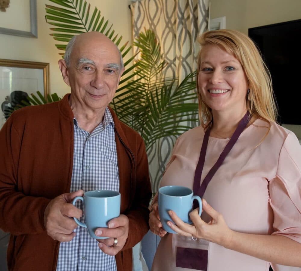 Older man with grey hair wearing a sweatshirt with his carer with long blonde hair both holding a cup of coffee and both standing inside the home smiling