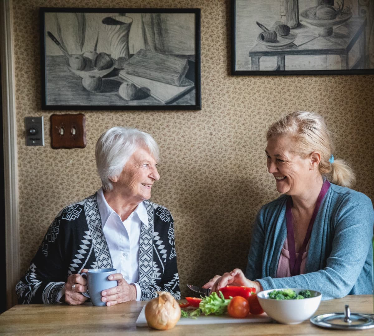 Older woman with grey hair chatting with her younger female carer while cutting vegetables both sitting on a chair inside the kitchen while chatting