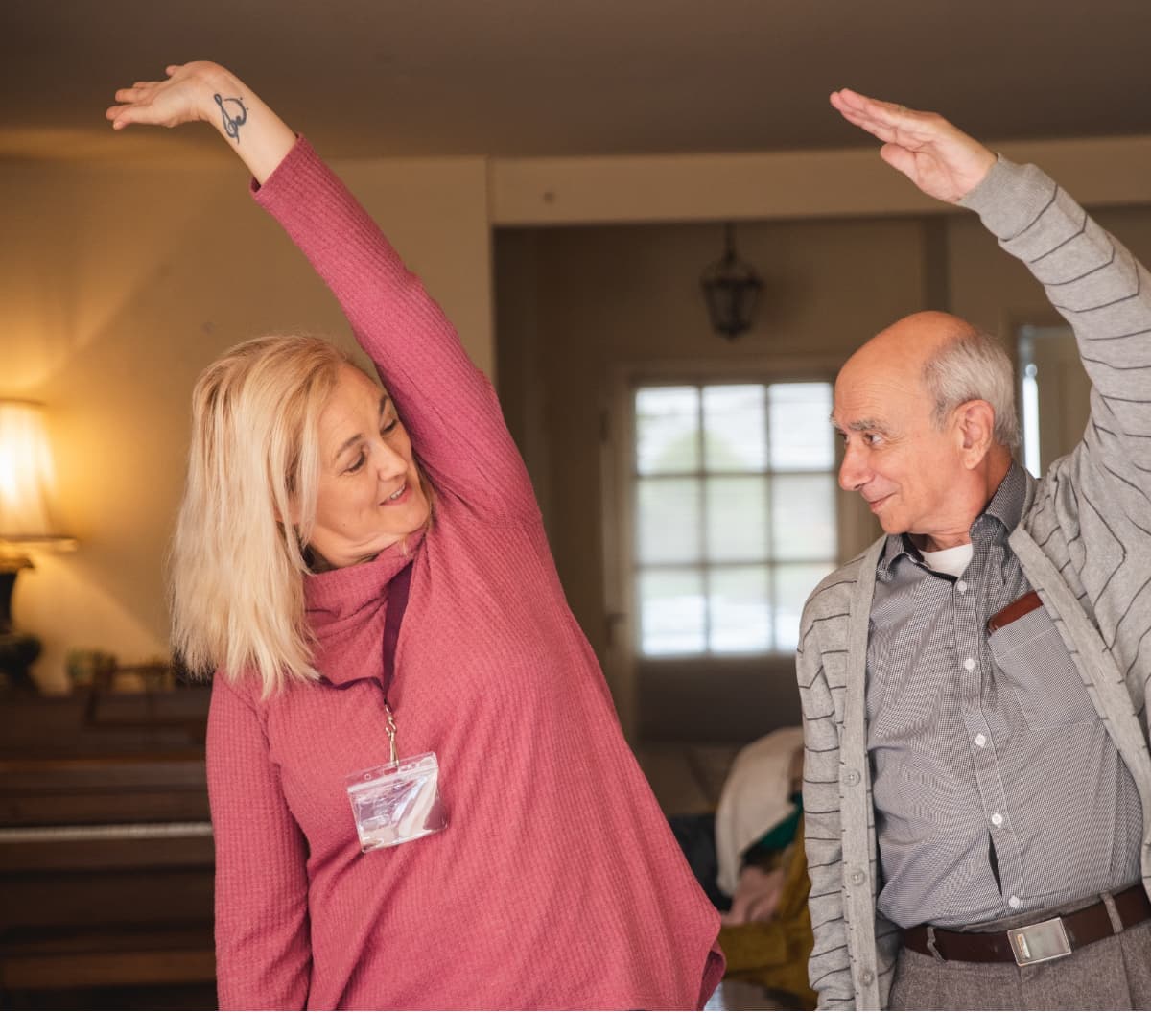 Older man with grey hair inside his house exercising with his younger female carer with blonde hair and wearing pink top