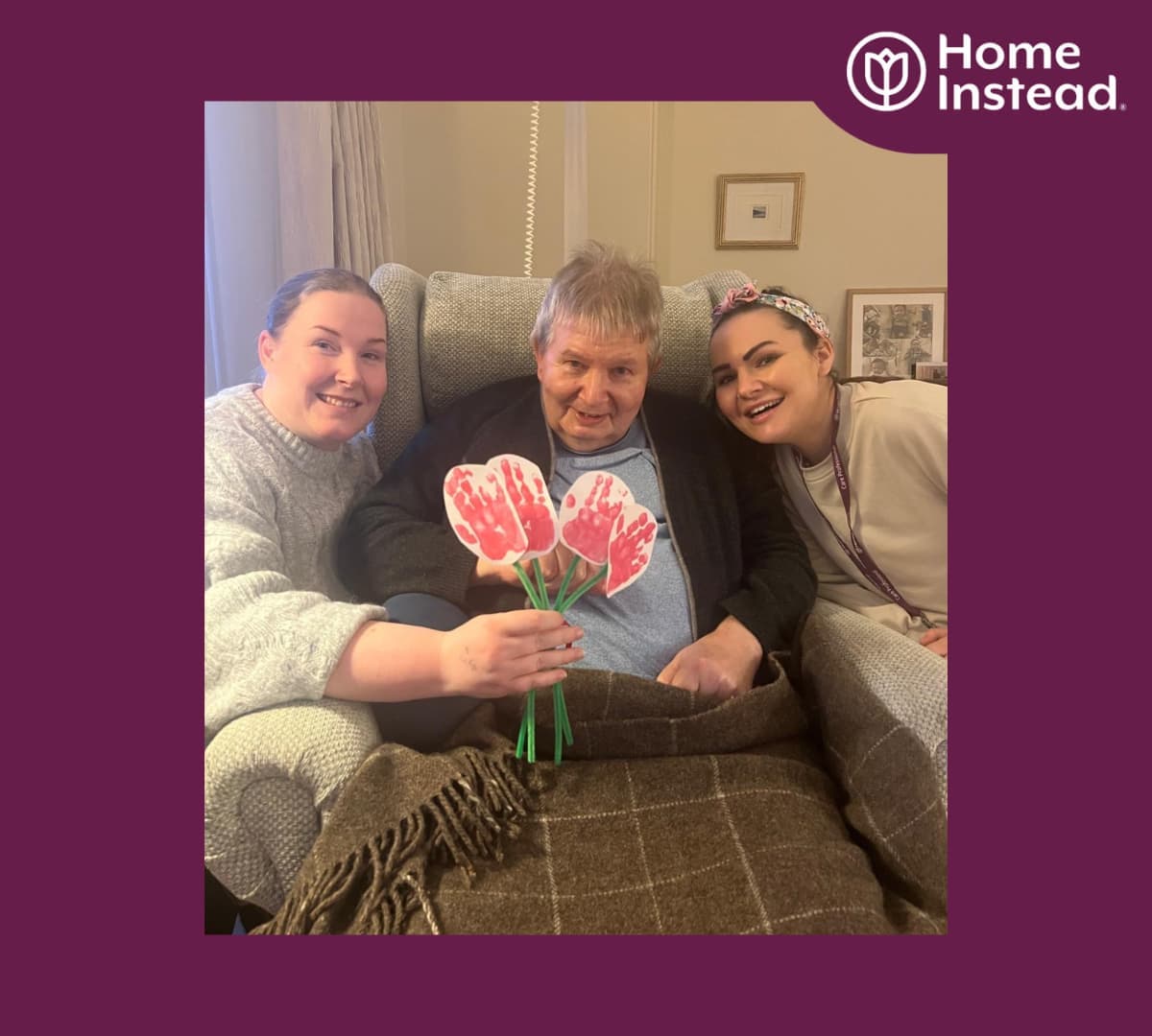 Two women and an older man holding a paper flower while sitijng on a couch, all smiling and happy