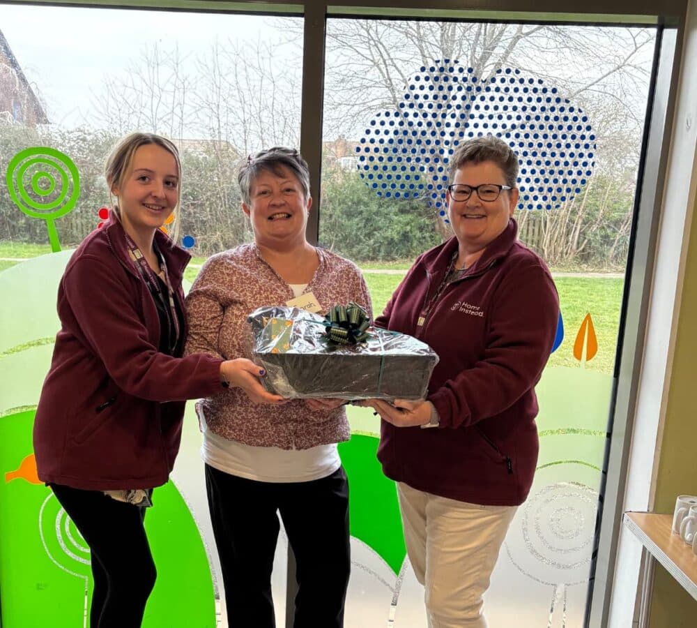 Woman holding a hamper inside the office with glass doors at her back and with two women helping her carry the basket all happy and smiling