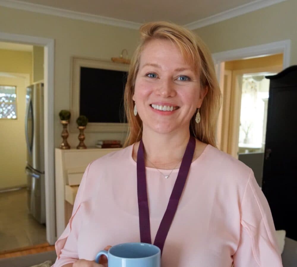 A woman carer with long blonde hair smiling wearing pink and holding a blue mug inside the home