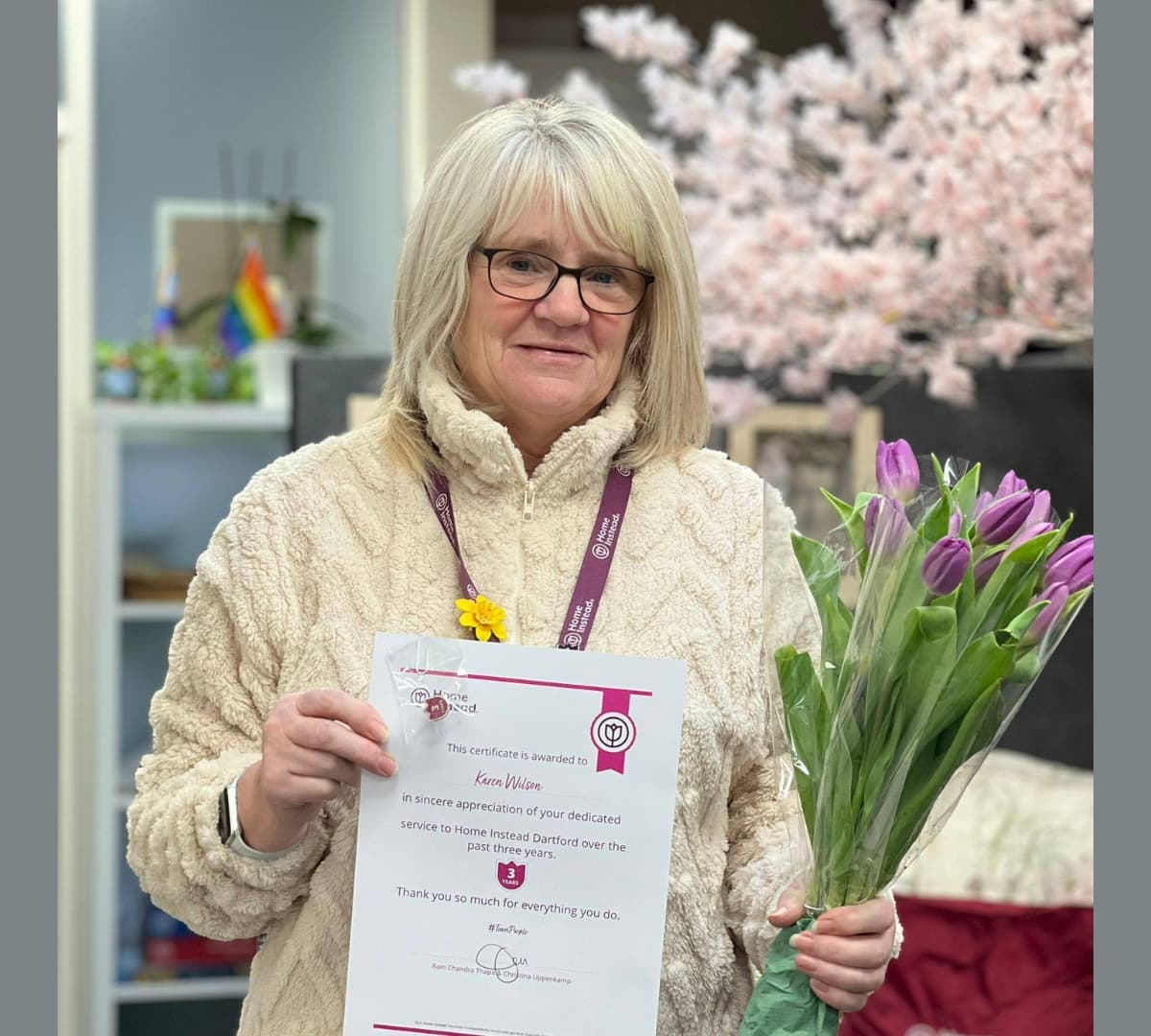 A woman receiving an award smiling while wearing eyeglasses and holding a certificate and some flowers