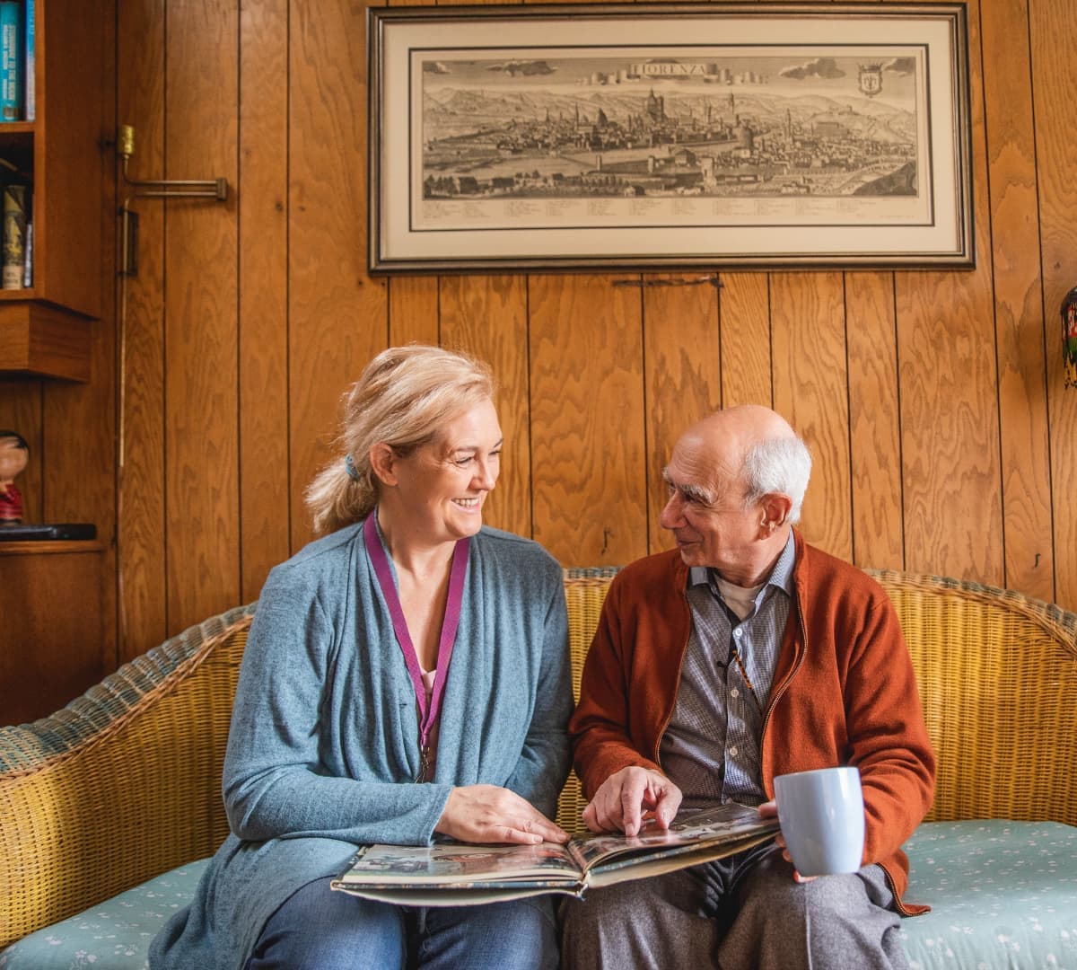 Older man with grey hair sitting on a couch with his younger female carer while looking at a photo album inside the home