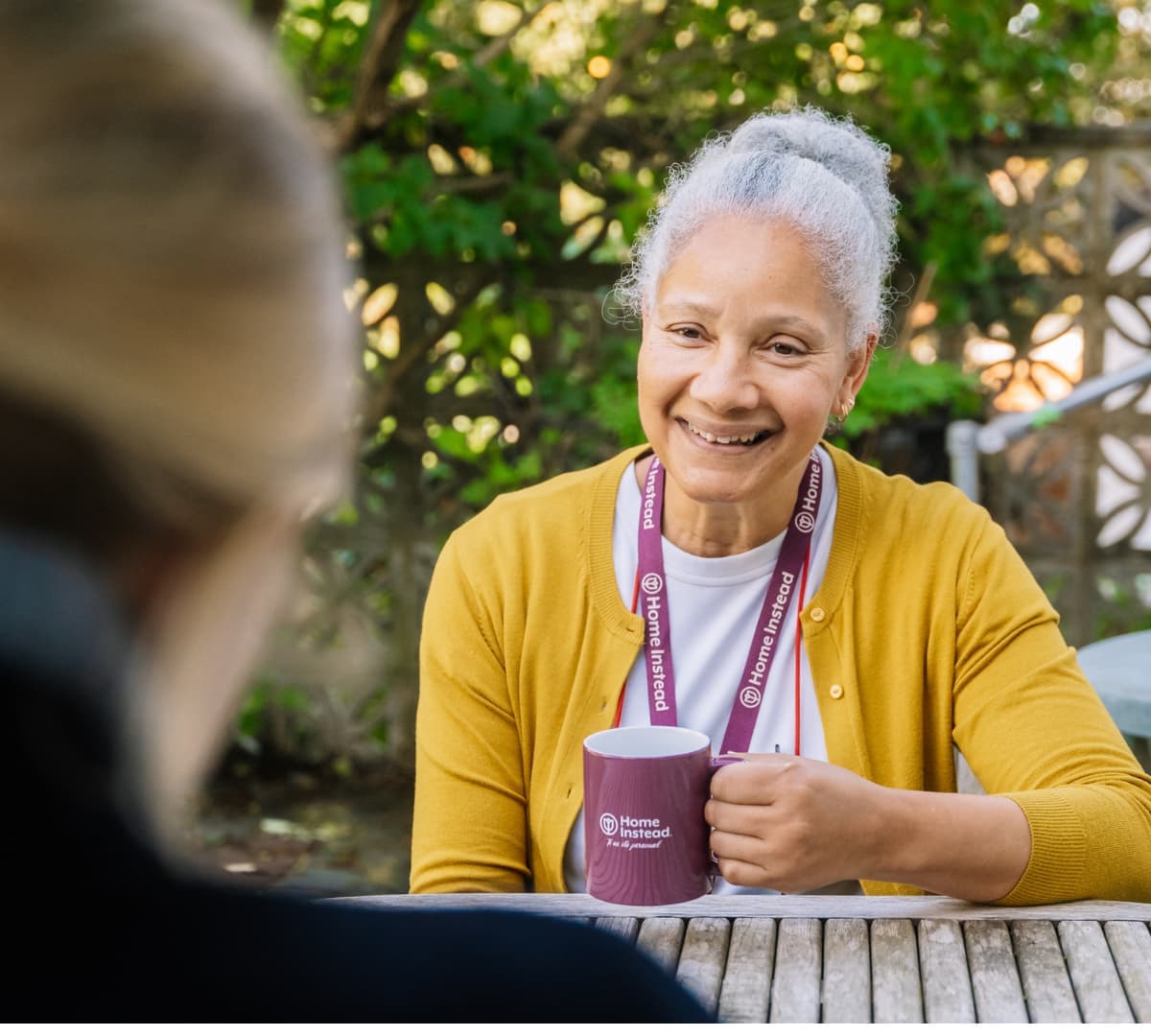 Older woman with grey hair and wearing a yellow sweater sitting on a chair outdoors while speaking to someone and holding a Home Instead mug