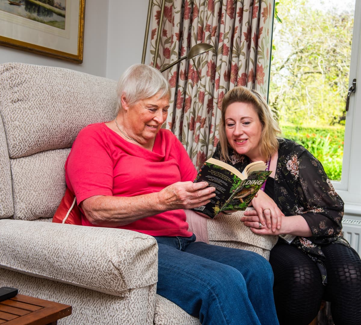 An older adult sitting on a couch while reading a book with her younger female carer