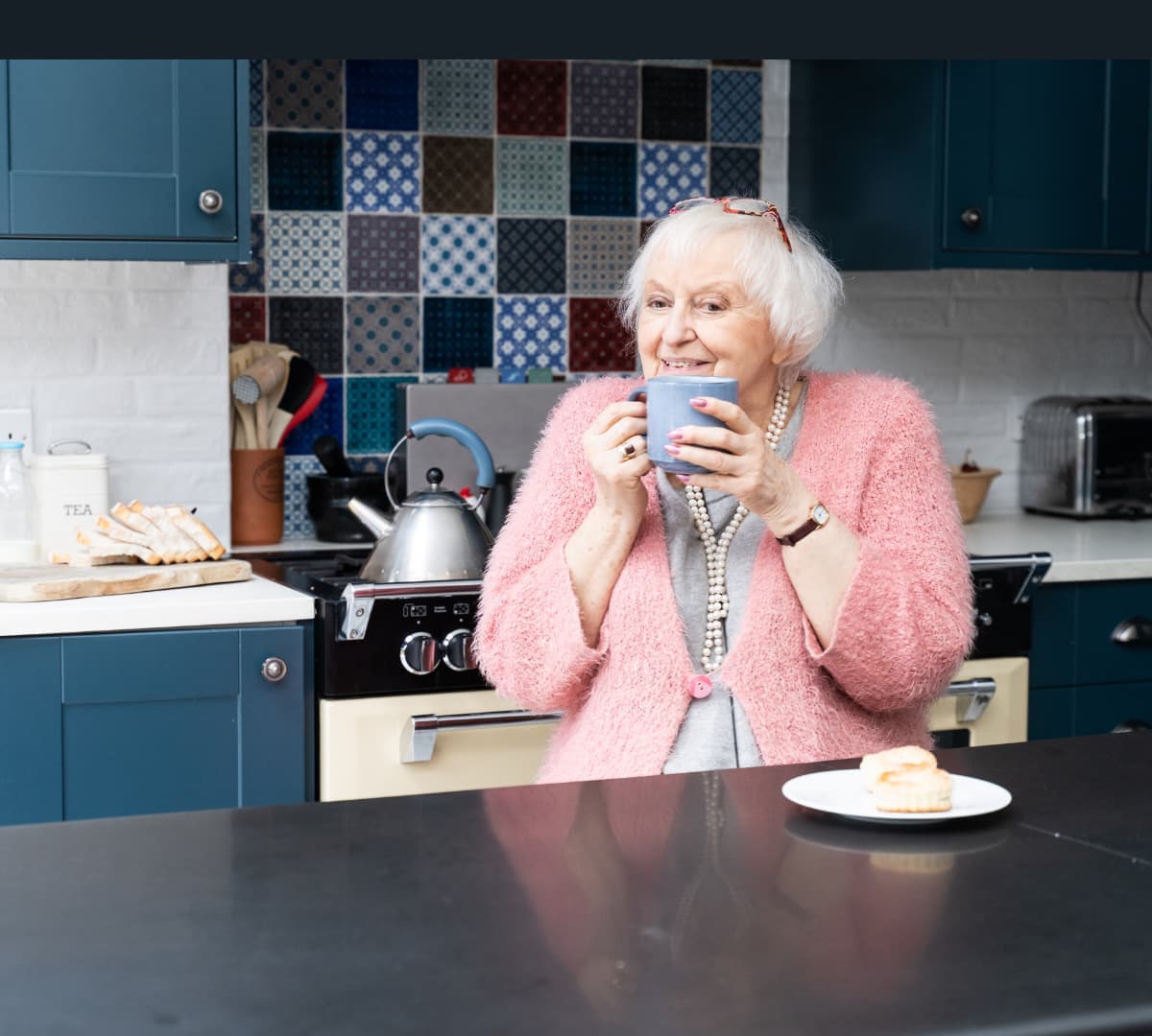 An old woman with white hair sipping coffee while wearing a pink sweater inside her kitchen
