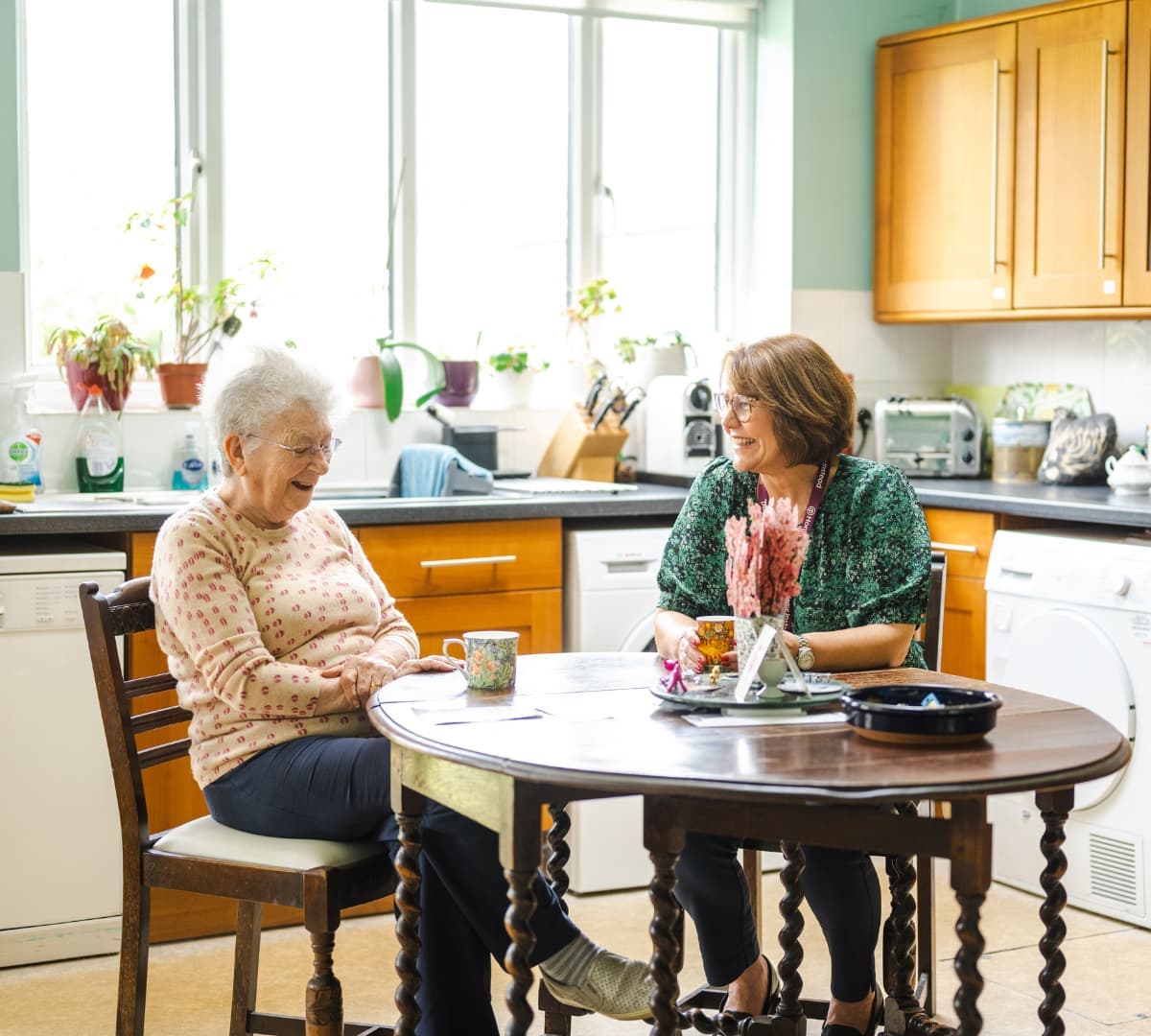 Two women chatting inside the kitchen while sitting on a chair both happy and smiling