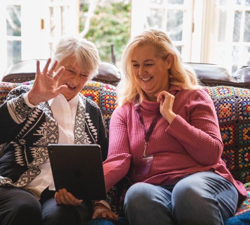 Two women talking to someone using an IPAD inside the house and near the window while sitting on a couch