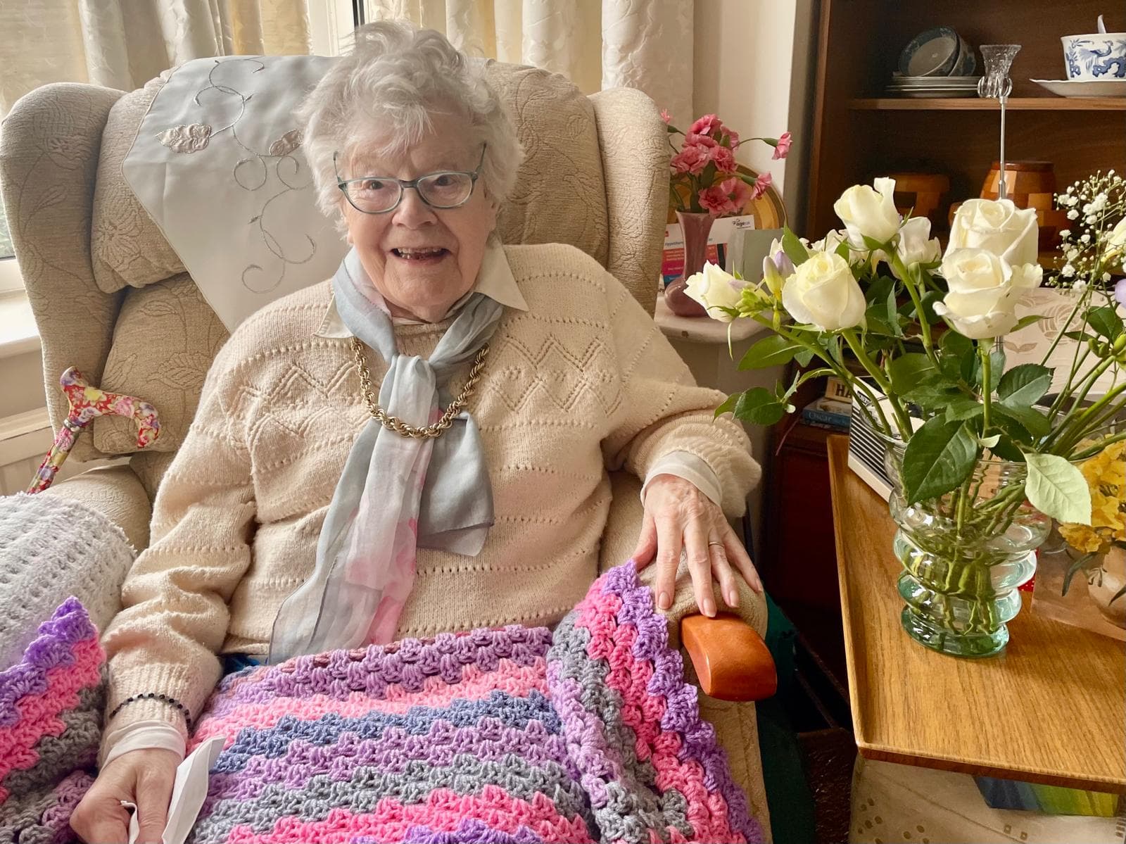 Smiling elderly woman with glasses sits in an armchair, holding a colorful knitted blanket next to white roses. - Home Instead