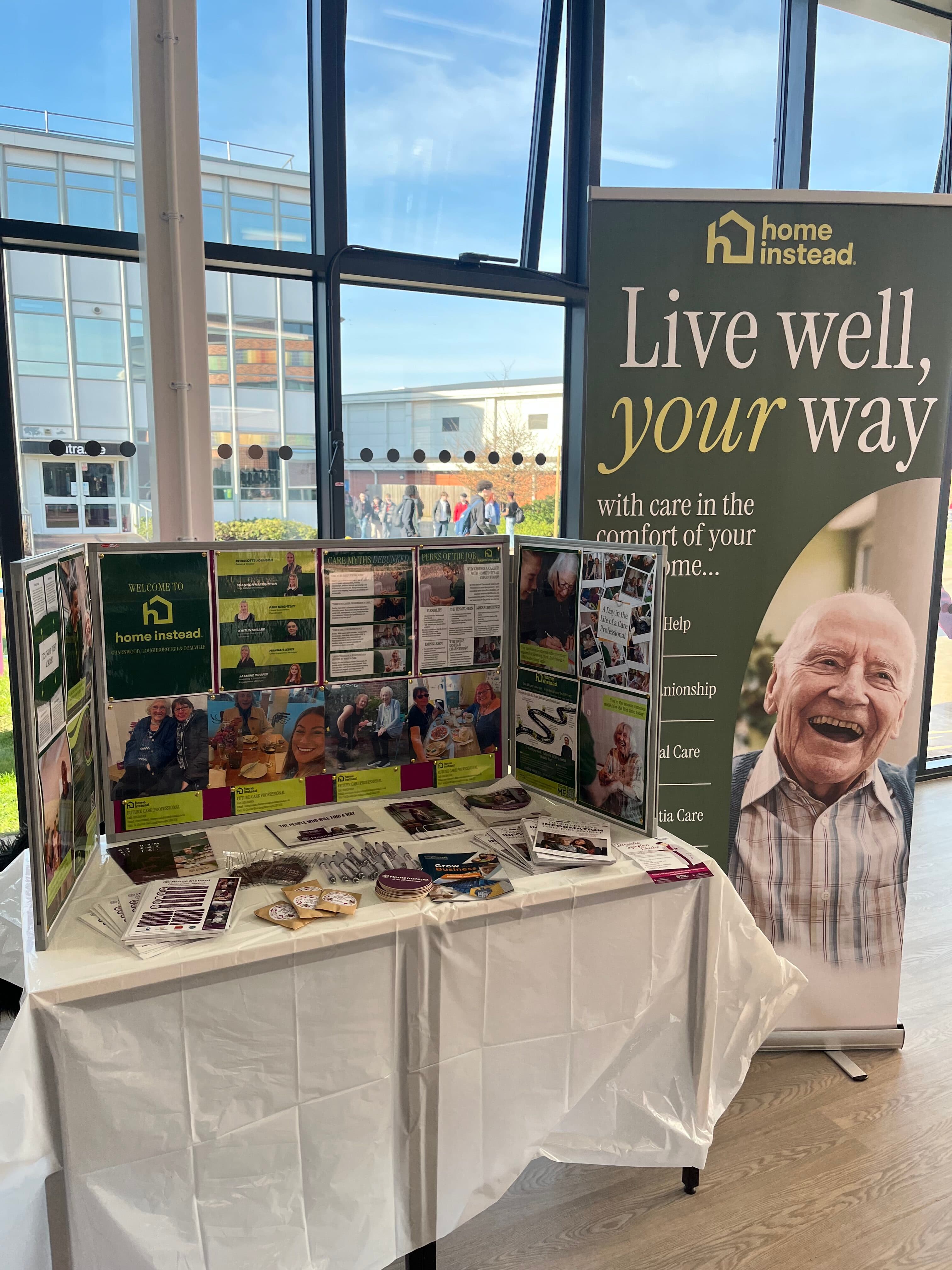 A display table for Home Instead with brochures, photos, and a banner showing a smiling elderly man. - Home Instead