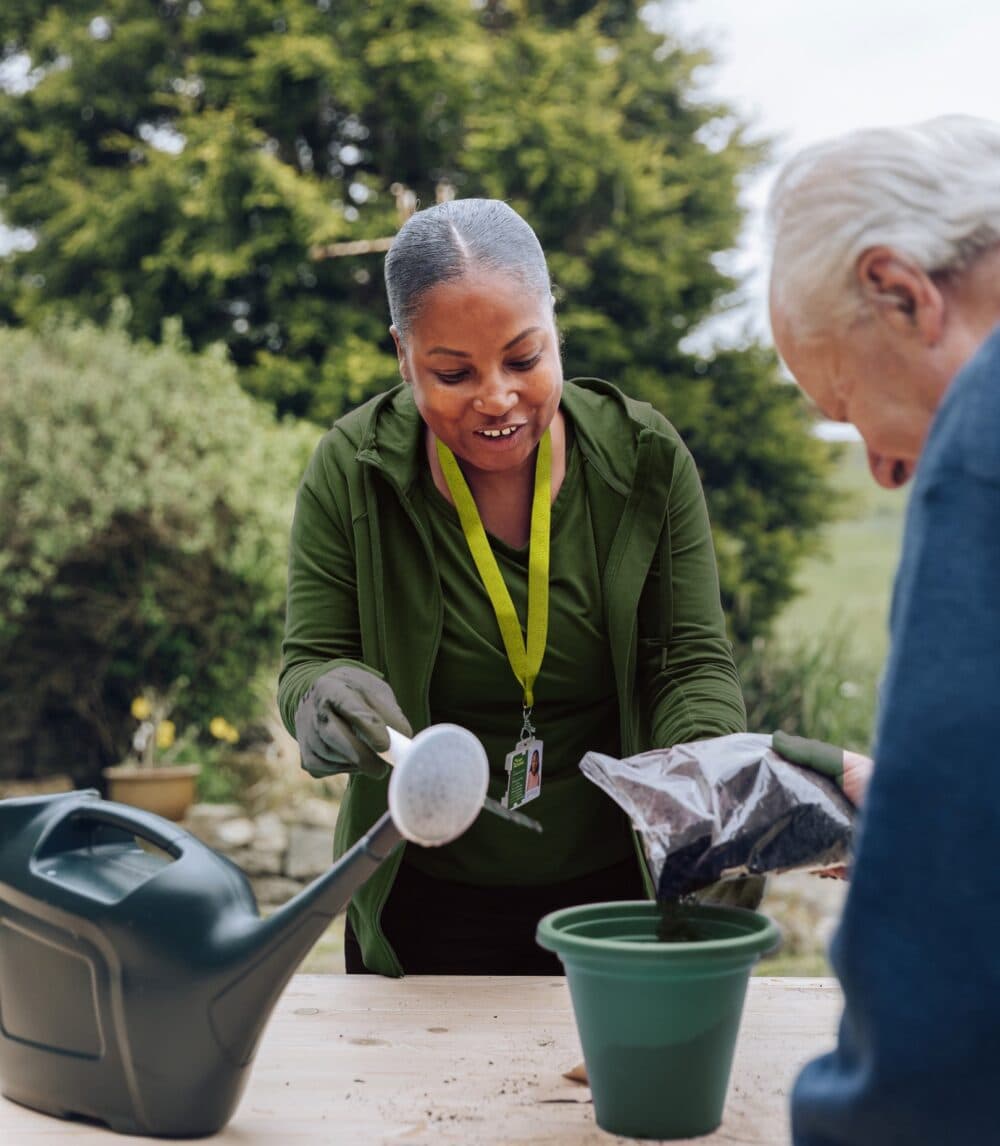 Smiling woman helps an elderly man pot a plant outdoors, holding soil beside a green watering can. - Home Instead