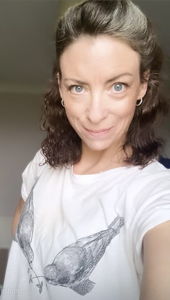 Woman with wavy brown hair smiling, wearing a white shirt with two illustrated birds facing each other. - Home Instead