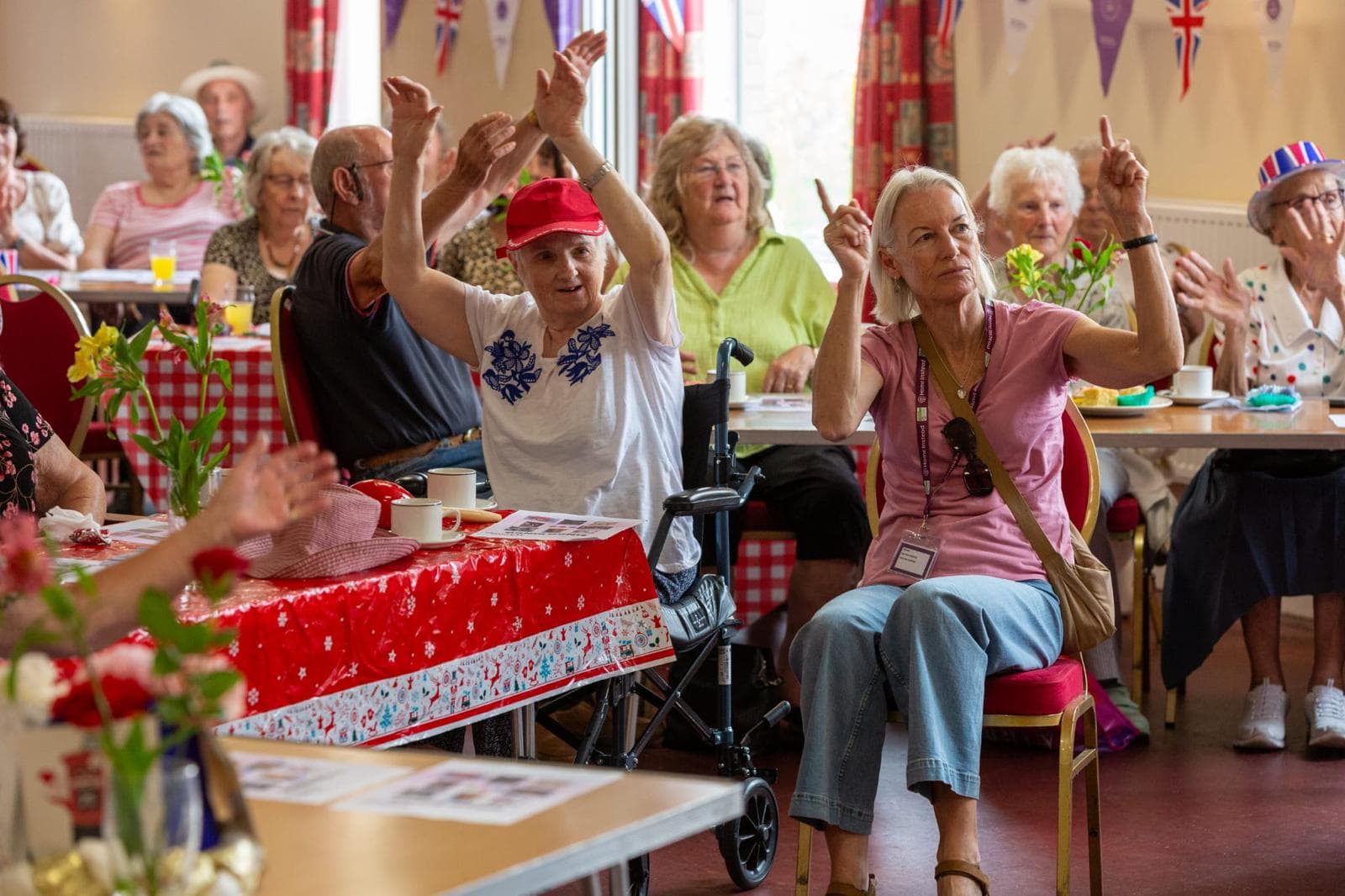 Elderly people at a decorated event, clapping and raising hands while seated around tables. - Home Instead