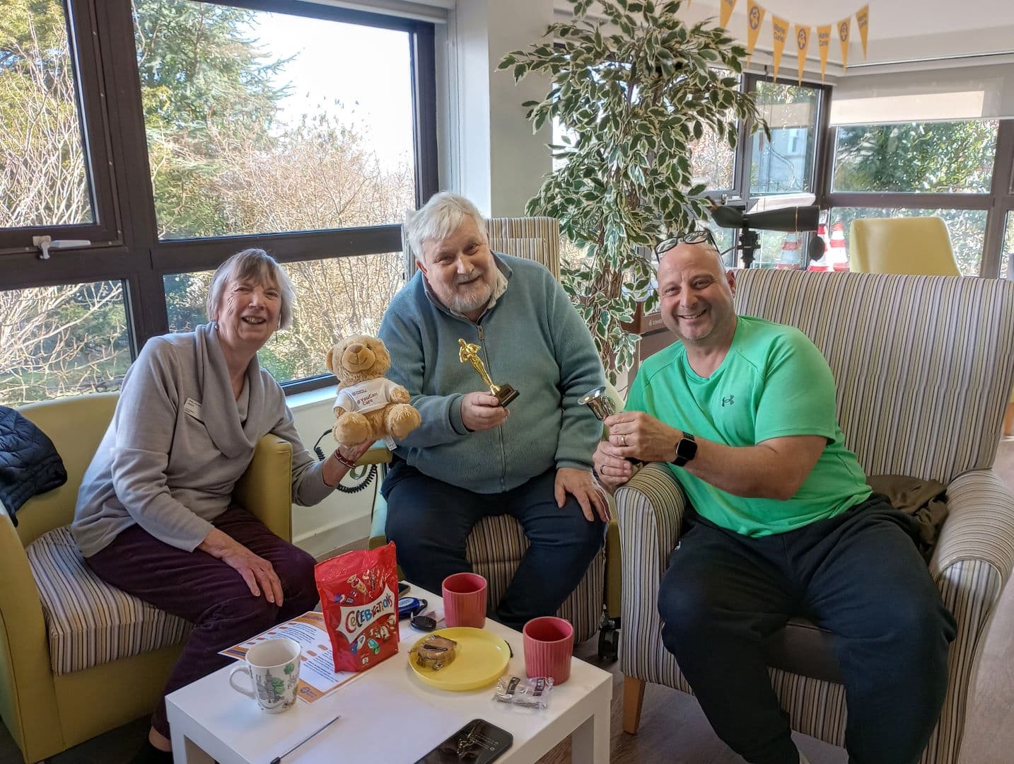 Three smiling adults sitting indoors, holding trophies and a teddy bear, with snacks and drinks on the table. - Home Instead