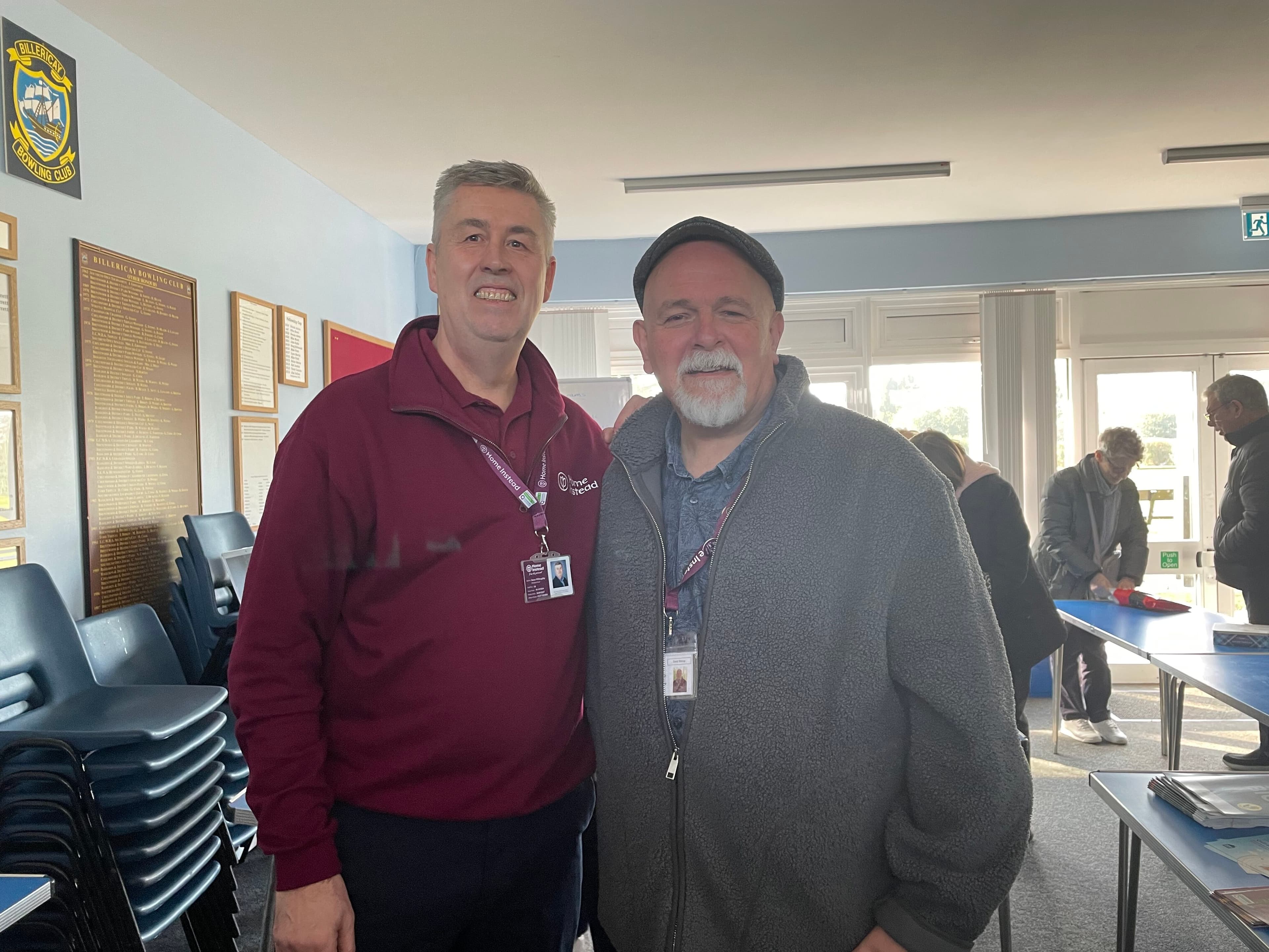 Two smiling men pose together in a bright room with stacked chairs and people in the background. - Home Instead