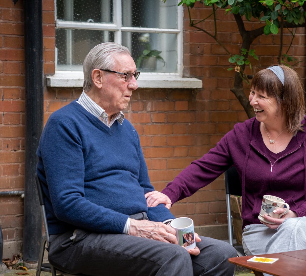 A young carer with short hair and purple sweater sitting and holding a cup of coffee outside the house with an older male adult with grey hair and wearing eyeglasses and blue sweatshirt both happy and smiling