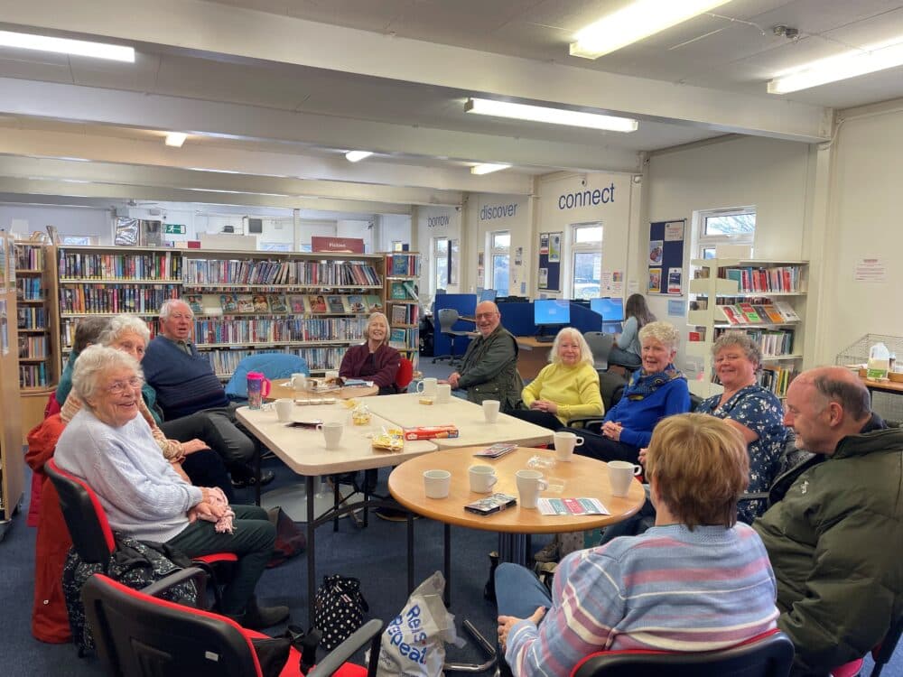 A group of older adults sit in a library around tables with drinks, snacks, and books, smiling and talking. - Home Instead
