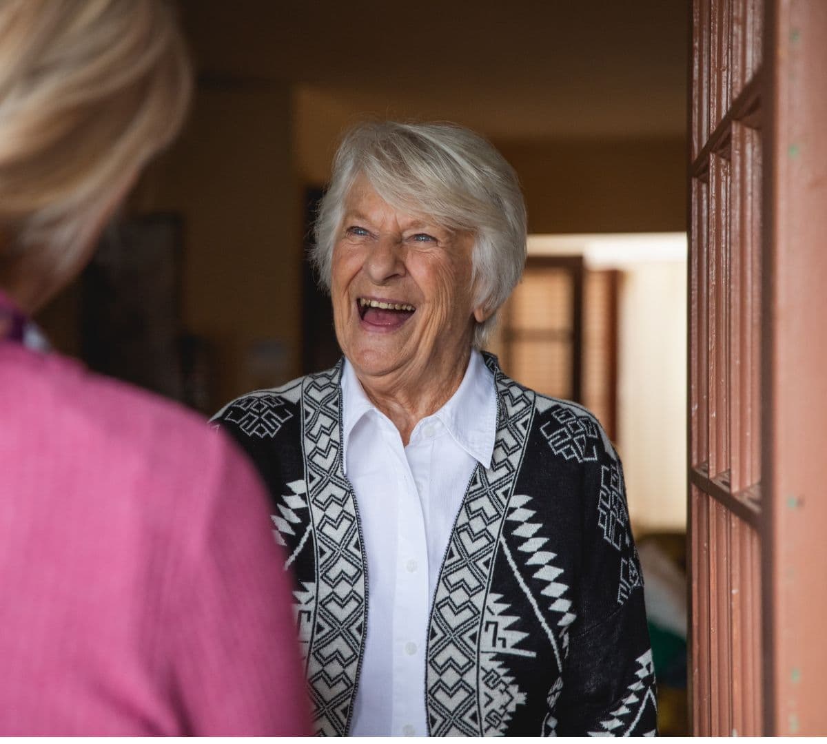 Older woman at the door wearing black printed top and with grey hair super happy while meeting her carer at the door
